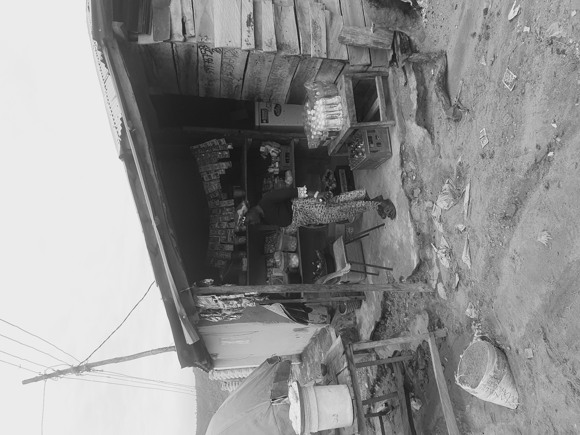 The image depicts a small, informal retail shop in Lokoja, Nigeria. The shop is constructed of wood and corrugated metal sheets. A woman is sitting inside, presumably the shopkeeper. Various items for sale, including bottled water, snacks, and drinks, are displayed on shelves. Outside the shop, there are plastic buckets, a wooden structure, and a bag. The surrounding area appears to be a dirt road with scattered debris. Overhead, there are utility poles and electrical wires.