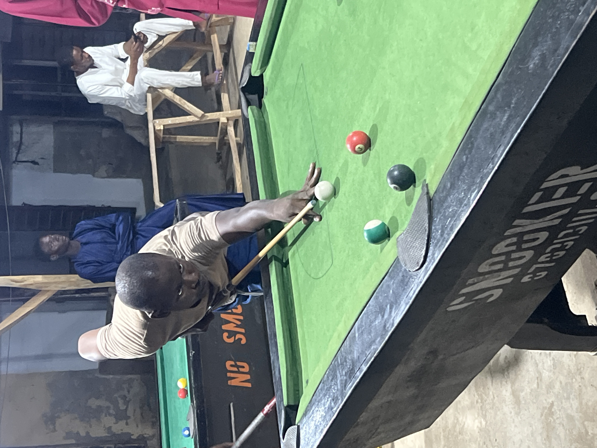 This vertical-oriented photograph captures an indoor scene, likely a recreational hall in Minna, Nigeria, where men are engaged in a game of snooker or pool. The central figure is a dark-skinned man, dressed in a light brown t-shirt, leaning over a green-felt snooker table. He is focused on making a shot, holding a wooden cue stick with his left hand forming a bridge over the table to guide the cue towards the white cue ball.
On the table, near the player's hands, are a white cue ball, a red ball, a black ball, and a green-and-white striped ball. Other colored balls are visible further down the table. In the background, two other dark-skinned men are present. One man, wearing a blue garment, stands or sits casually, observing the game. To his left, another man in white traditional attire is seated on a wooden structure, preoccupied with his mobile phone.
The setting appears to be a casual, somewhat dimly lit space, suggesting it might be evening or night. The background features dark walls and what looks like wooden scaffolding or improvised seating. Visible text on the side of the snooker table includes "SNOOKER" in white letters, and partially visible text that reads "NO SM..." (likely part of "NO SMOKING"). Some indistinct numbers, possibly "G763131," are also seen on the table's side. The overall scene depicts a common leisure activity in a local context.