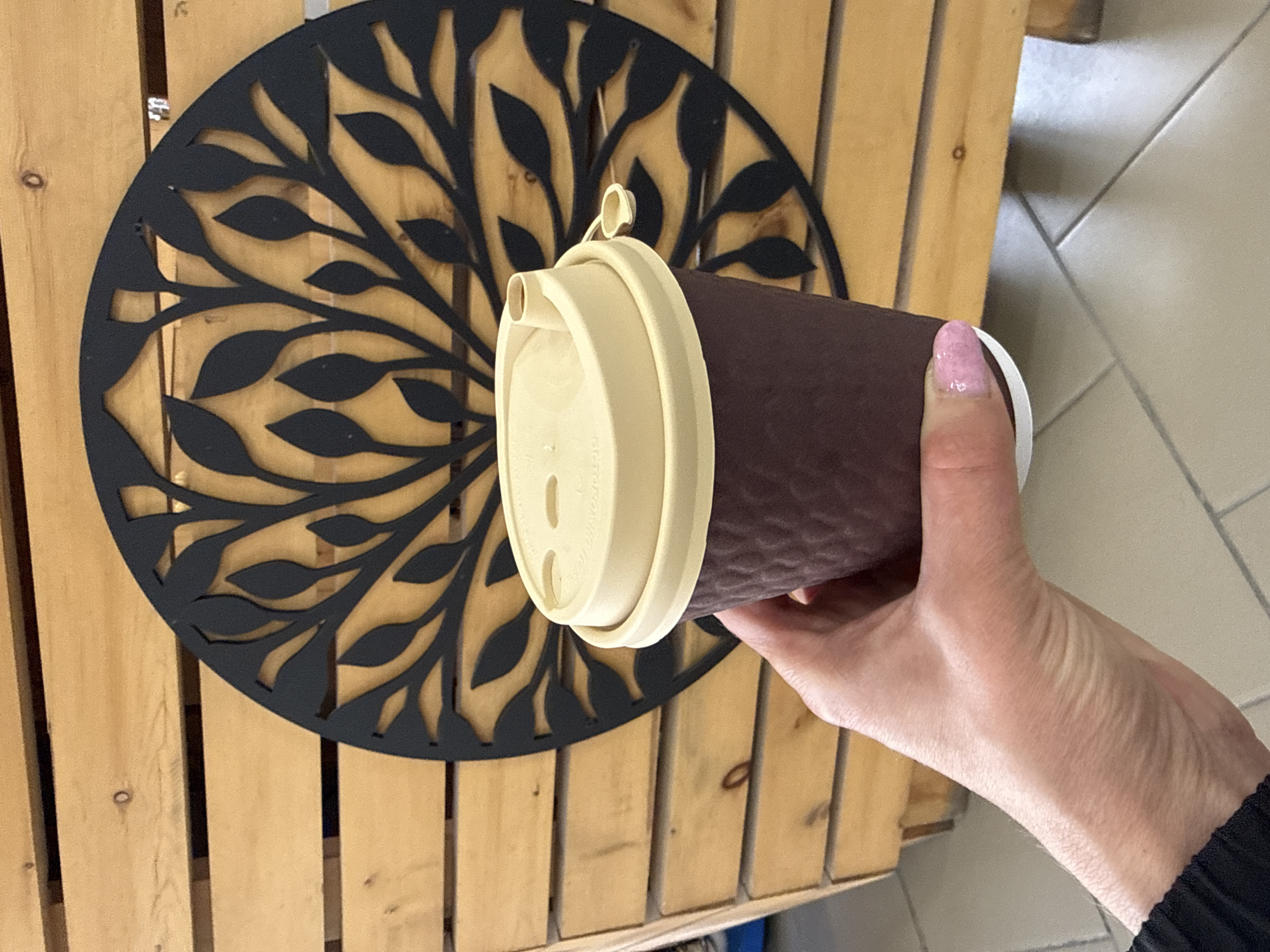 A hand holds a brown, textured coffee cup with a beige lid. The background features a wooden surface with a circular, black, leafy-patterned decoration. The image appears to have been taken in an indoor setting, possibly a café, as evidenced by the wood paneling and tiled flooring visible in the periphery. The location is Ust-Koksa, Russia.