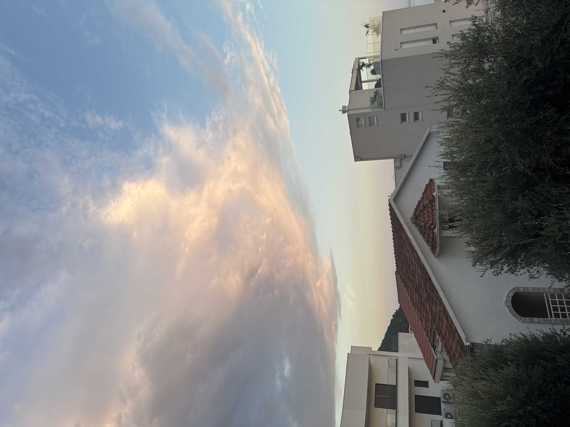 This image depicts a view of buildings and the sky in the city of Trap, Montenegro. The foreground is dominated by the dense, dark green foliage of trees, likely olive trees given the Mediterranean setting.
In the mid-ground, several buildings are visible. To the left, a portion of a modern, multi-story white building with balconies and dark-framed windows is present. Adjacent to this, a smaller white building with a traditional red-tiled roof stands out. This building features an arched entrance with stone accents and a white railing. To the right, a taller, contemporary white building with multiple windows and a rooftop terrace with a glass railing and some potted plants is situated. The sea is faintly visible in the far right background.
The sky above is a prominent feature, displaying a mix of soft pastel colors indicative of sunset or sunrise. Large, wispy clouds in shades of pink, orange, and pale blue stretch across the frame, contrasting with areas of clear blue sky. The lighting suggests it is either early morning or late evening. There are no people clearly visible in the image.