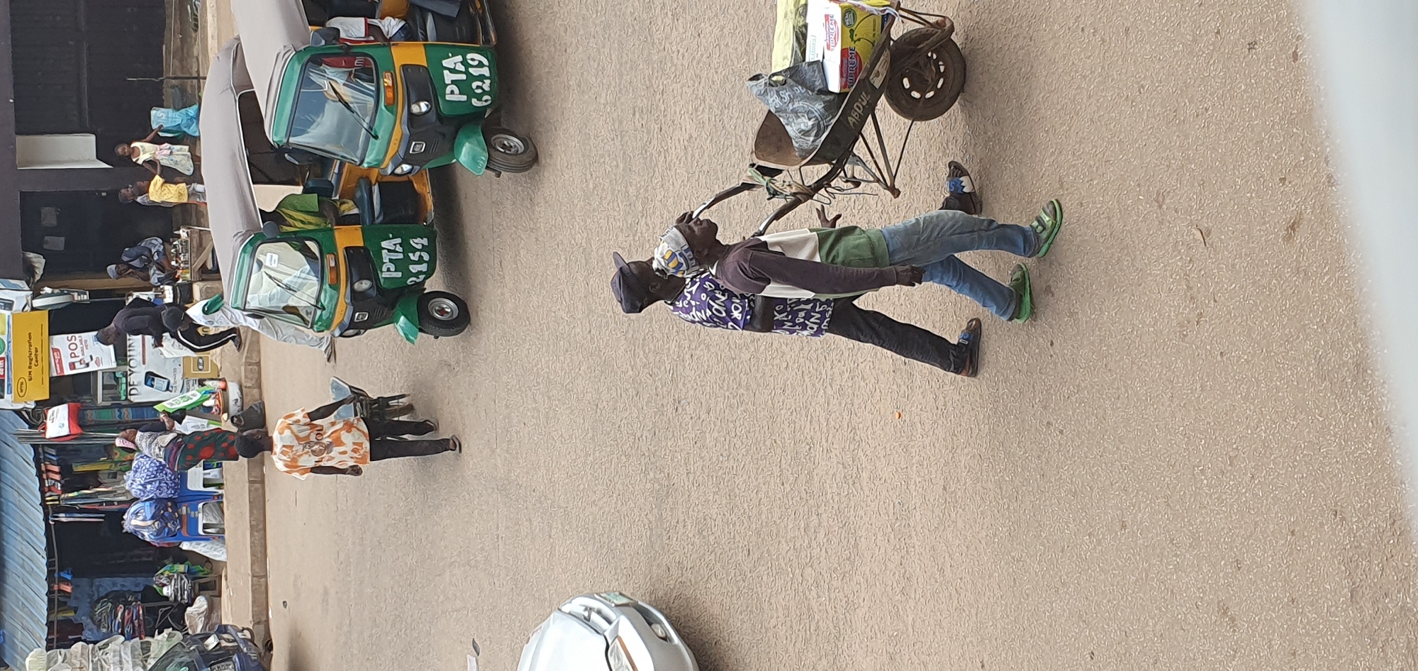 In Bukuru, Nigeria, a street scene captures a moment with several individuals and vehicles. In the foreground, two men are pushing a wheelbarrow loaded with goods. The man in front, wearing a patterned shirt and a cap, is pushing the wheelbarrow. Behind him, another man in a brown and green long-sleeved shirt and blue jeans is also assisting. The wheelbarrow contains various items, including what appears to be bags and a yellow box labeled "SUPREME". The men are walking on a paved surface that appears to be a street.
In the middle ground, two green and yellow three-wheeled auto-rickshaws are parked, one behind the other. The front auto-rickshaw displays the license plate "PTA 2154", and the rear one has "PTA 6219". A person is visible in the driver's seat of the front auto-rickshaw.
Further in the background, a street market or shop area is visible. Stalls are set up with various goods displayed, and several people are interacting within this area. One person in an orange and white patterned shirt is walking away from the camera, pushing a wheelbarrow, while other individuals are seen standing or sitting. A silver car is partially visible on the left side of the image. The overall environment suggests a busy urban setting with commercial activity.