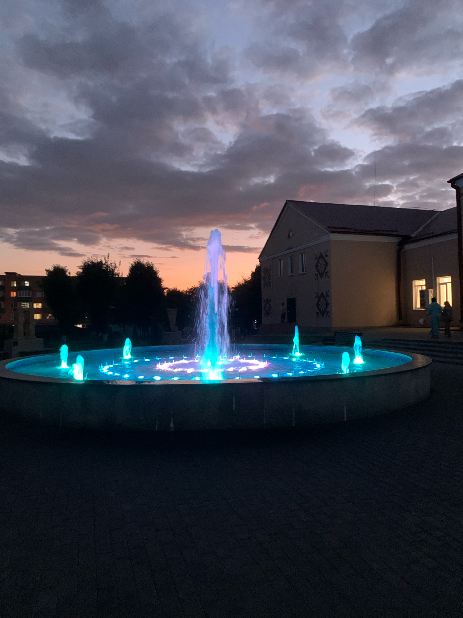 The image depicts an outdoor scene at dusk in Kapyĺ, Belarus, focusing on a circular fountain. The fountain is illuminated with vibrant blue and green lights, with water jets creating a dynamic display. The sky above is filled with dramatic, cloudy formations, tinged with the soft hues of sunset. In the background, a building with a light-colored facade and decorative elements is visible. To the left, a residential building with illuminated windows suggests a nearby urban area. The ground in the foreground is paved with dark stone or brick. The overall atmosphere is serene and visually striking due to the contrast between the natural light of the sky and the artificial illumination of the fountain.