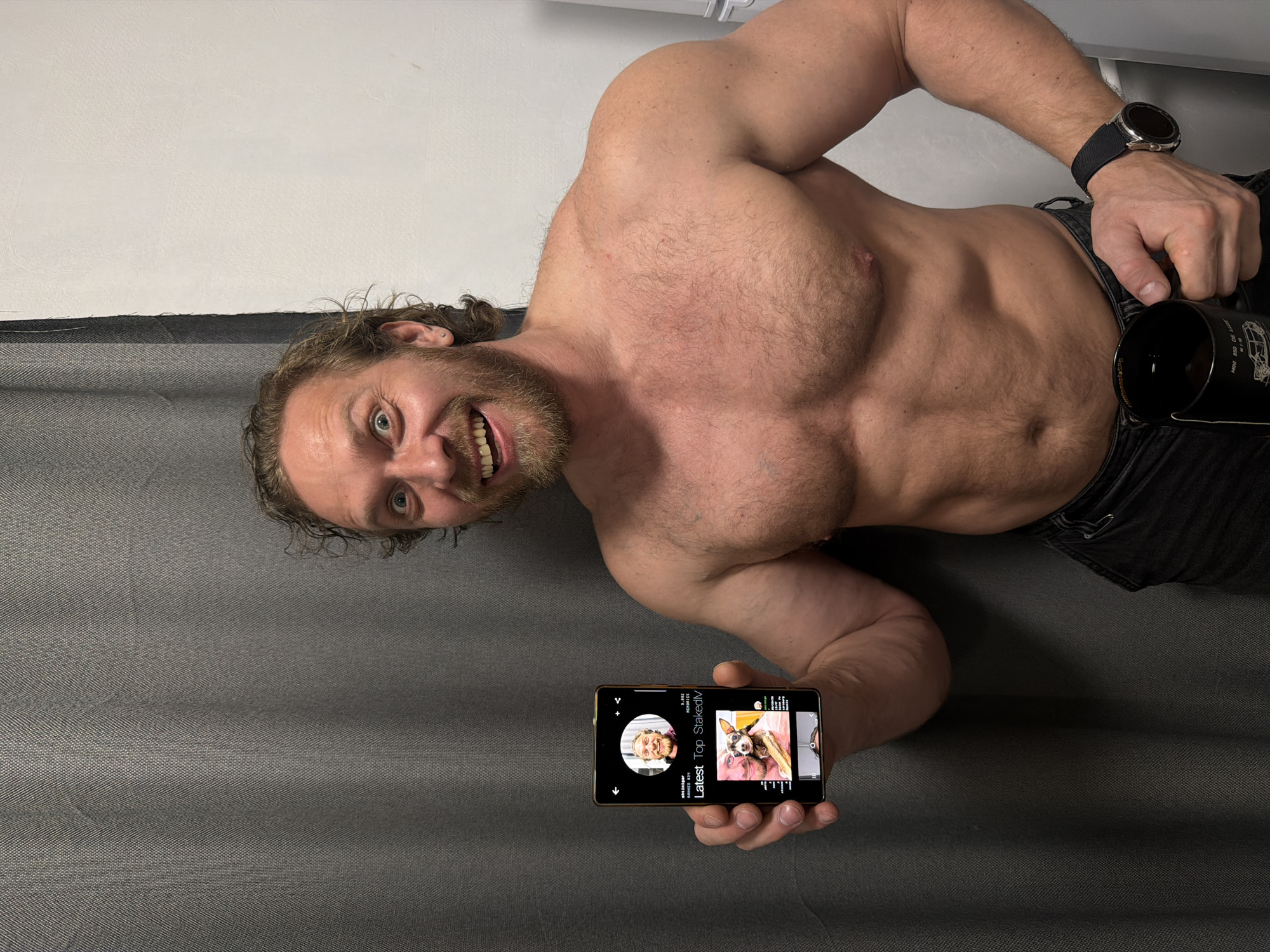 A shirtless, muscular man with long hair and a beard is lying on a gray surface. He is holding a smartphone in one hand and a black mug in the other. He is smiling and looking at the camera. He is wearing a watch and black pants. The background is a plain white wall. The location is not discernible from the image.