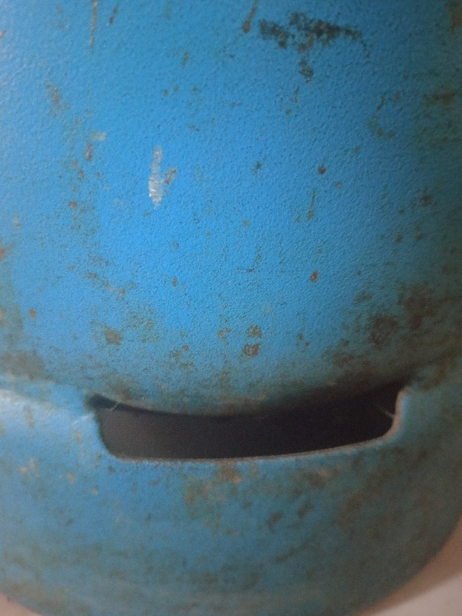 The image shows a close-up, abstract view of a weathered blue surface with visible rust spots and a dark, curved opening. The texture of the surface is rough and uneven, suggesting age and exposure to the elements. There are no discernible people, specific objects, or actions taking place within the frame. The focus is entirely on the material and its deterioration. The location in Mukapayung, Indonesia, is not visually represented in the image itself.