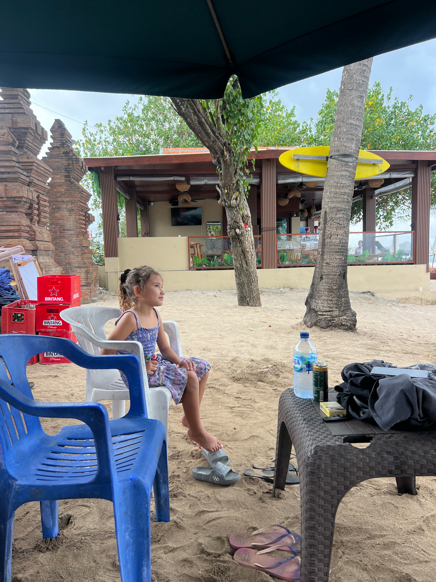 The image depicts a beach scene in Kuta, Indonesia. A young girl with curly hair, wearing a purple floral dress, sits on a white plastic chair, looking to her right. In the foreground, a blue plastic chair faces the viewer. To the right of the girl, a brown woven table holds a bottle of water, a can, a pack of cigarettes, and folded black clothing. A pair of flip-flops rests on the sand near the table. Behind the chairs and table, a beach restaurant with wooden beams and a thatched roof is visible. A yellow surfboard is attached to a palm tree near the restaurant. To the left, a brick structure resembling a temple gate stands against a cloudy sky. The ground is covered in sand.