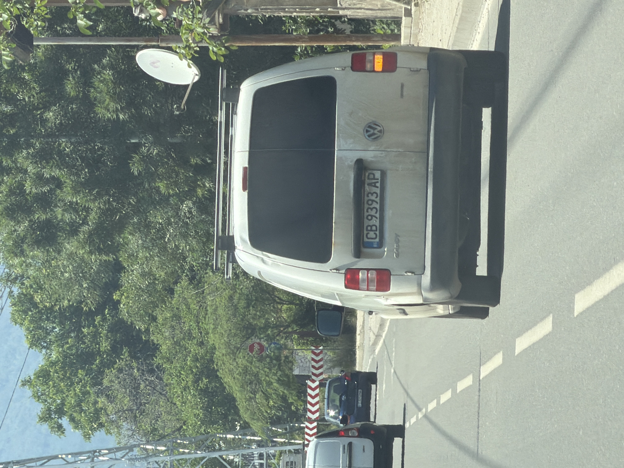 A white Volkswagen Caddy van with license plate CB 9393 AP is traveling on a paved road. The van is dirty and has a roof rack. The license plate is located on the rear of the vehicle. The van is in the foreground, slightly to the right of the frame, with its rear end facing the camera. The vehicle is driving on a two-lane road with a dashed white line marking the lanes. In the background, there is a blue hatchback car and another white van, indicating traffic. A red and white chevron barrier is visible on the left side of the road, suggesting a potential road closure or construction. Further in the background, lush green trees form a dense foliage. A stop sign is posted on a pole amidst the trees. A satellite dish is mounted on a pole to the right of the van. The scene appears to be in a suburban or semi-rural area, possibly on the outskirts of Sofia, Bulgaria, given the Bulgarian license plate and the general environment. The lighting suggests it is daytime, with sunlight illuminating the scene. There are no people clearly visible in the image.