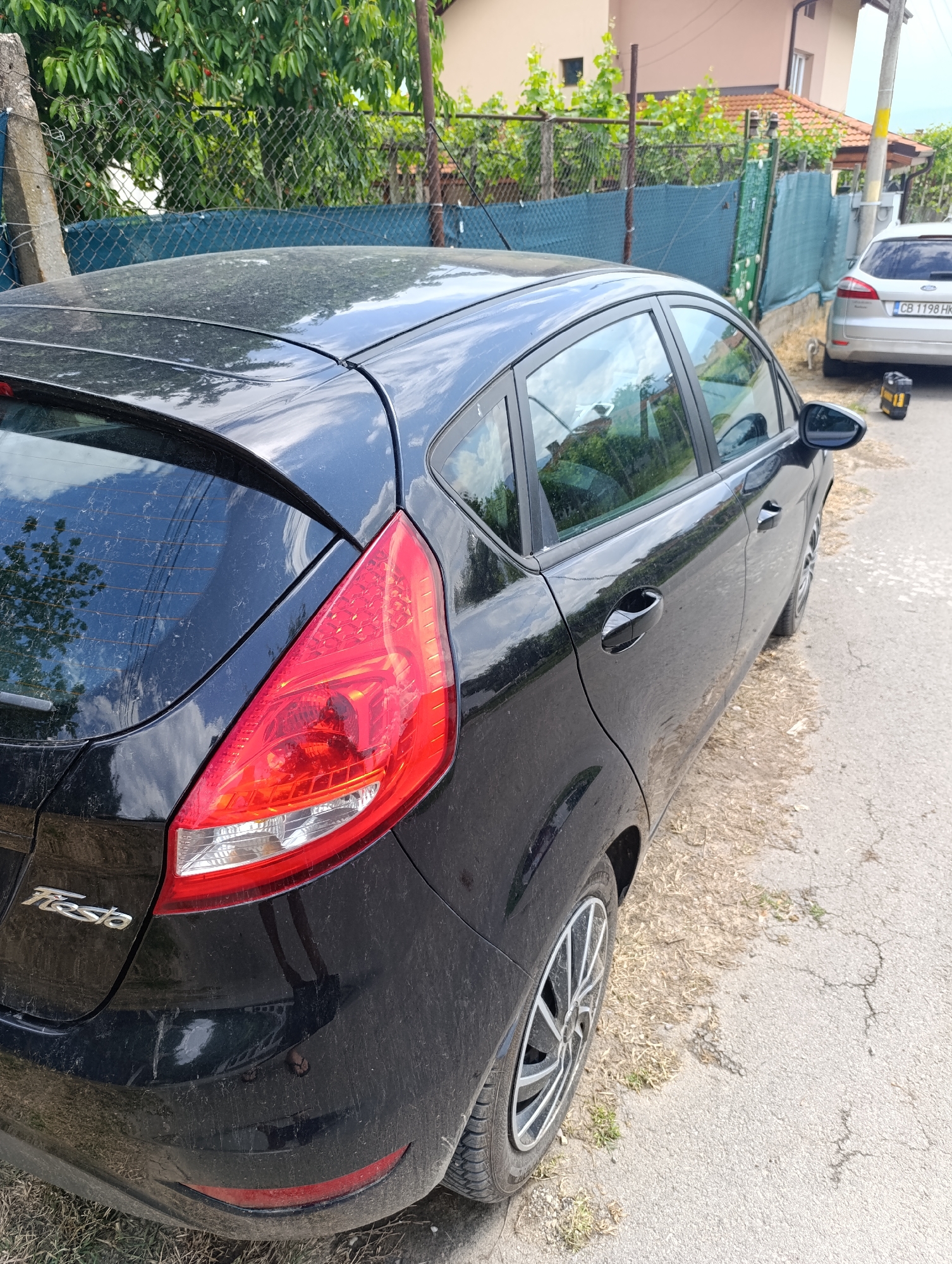 A black, heavily dust-covered Ford Fiesta hatchback is parked on an unpaved roadside in Nikolichevtsi, Bulgaria, viewed from its rear right quarter. The vehicle's paintwork, roof, and rear window are obscured by a thick layer of fine dirt and water marks. The 'Fiesta' badge is affixed to the rear left. The right rear taillight is illuminated, and the right rear alloy wheel is visible. A light scuff is present on the lower left of the rear bumper. The road surface consists of cracked asphalt transitioning to a dirt and dry grass shoulder. In the background, a silver Ford wagon, bearing Bulgarian license plate 'CB 1198 HK', is parked further along the road, adjacent to a yellow and black container on the ground. A chain-link fence with blue privacy netting, backed by green deciduous trees showing red fruit, and a light-colored two-story building with a red tile roof, define the perimeter. A utility pole with yellow bands stands roadside. No persons are present.