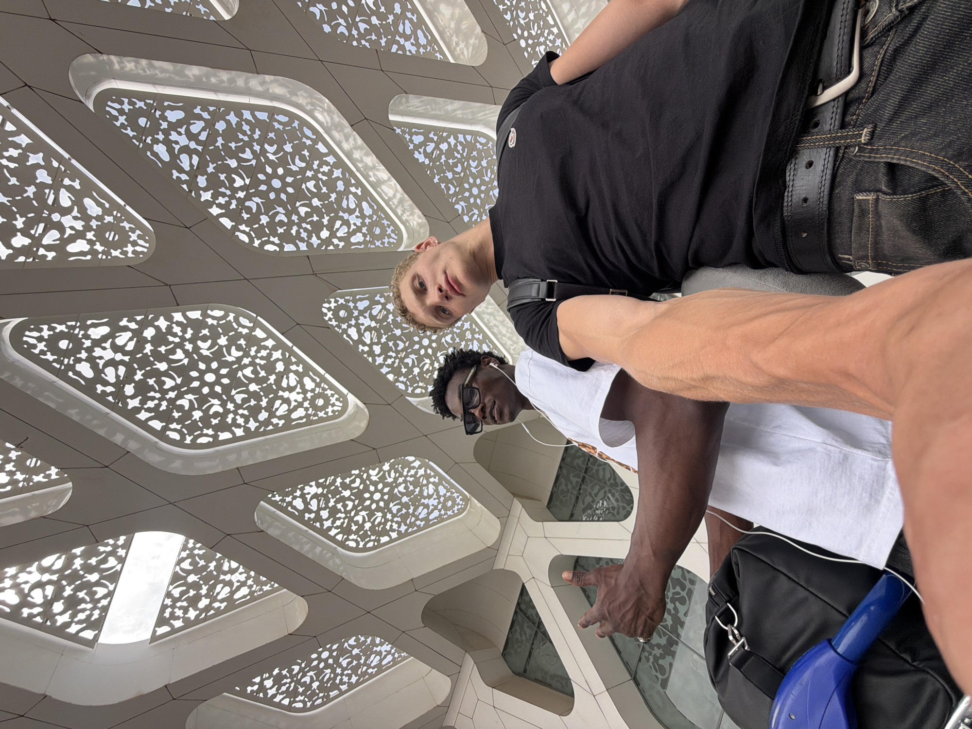 This is a low-angle, wide shot photograph taken from the perspective of a person holding the camera. Two young men are positioned under a modern, white, geometrically patterned roof structure, possibly a facade or canopy, with a bright sky visible through the openings. The man on the left, who is Black, is wearing a white t-shirt, sunglasses, and white earphones. His left arm is extended forward, and his hand is making a "shaka" gesture. He is also wearing a ring on his left hand. A black duffel bag and a blue item are visible near him. The man on the right, who is white, is wearing a black t-shirt, a backpack, and dark denim jeans with a black belt. His right arm is in the foreground of the image. The architectural style suggests the image was taken at Marrakech Menara Airport in Morocco, known for its distinctive roof design.