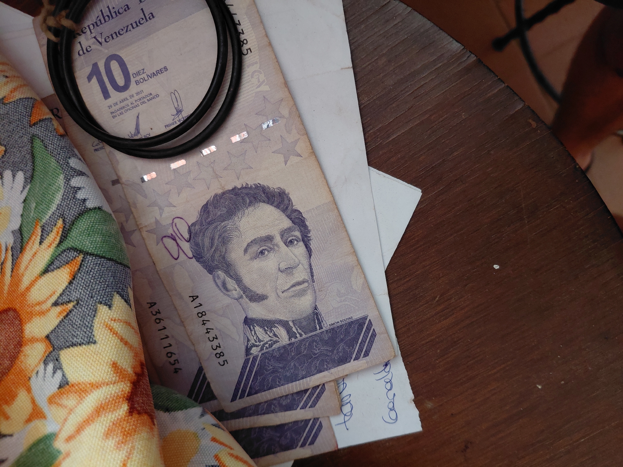 The image displays a close-up, overhead view of currency, fabric, paper, and a coiled black object resting on a dark brown wooden surface.
In the upper left quadrant, a segment of textured fabric with a sunflower pattern is visible. The pattern consists of yellow, orange, and white petals with green leaf accents against a grey background.
Beneath the fabric, several Venezuelan banknotes of 10 Bolívares are arranged. The most prominent banknote features a portrait of Simón Bolívar on its left side, with "SIMÓN BOLÍVAR" printed vertically along a purple band. The note displays "REPÚBLICA BOLIVARIANA DE VENEZUELA," the denomination "10 DIEZ BOLÍVARES," and the date "29 DE ABRIL DE 2021." Security features include metallic holographic stars. Visible serial numbers on different banknotes include "A18443385," "A3611654" (partially obscured), and "58227" (partially obscured).
Resting on top of the banknotes, in the upper right, is a black, flexible, circular object, possibly a coiled cable or bracelet.
In the lower left, a piece of white paper is positioned beneath the banknotes, showing handwritten text in blue ink. The visible words are "tama" (partially) and "Geraldo."
The entire arrangement is situated on a dark brown wooden surface, likely a tabletop, exhibiting visible wood grain.