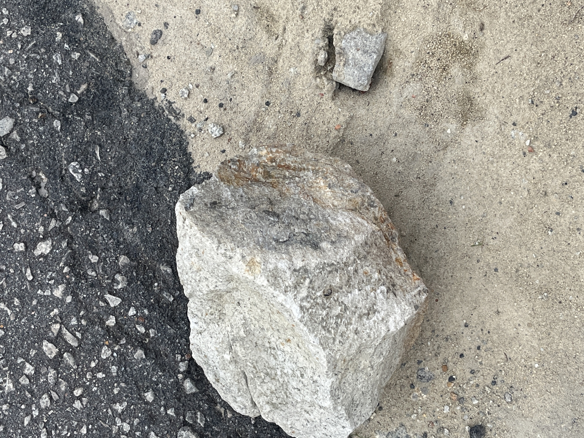 The image shows a close-up, overhead view of a large, light-colored boulder resting on a surface of sand and small gravel. To the upper left, a section of asphalt pavement with visible aggregate is visible. The transition between the asphalt and the sandy surface is clearly defined. The lighting suggests an outdoor, daylight setting. No people are present in the image. The scene depicts a natural or urban debris situation, possibly indicative of construction, roadwork, or a natural event. The specific location in Teluk Air, Indonesia, is not visually evident from the image itself, but the description is provided as requested.