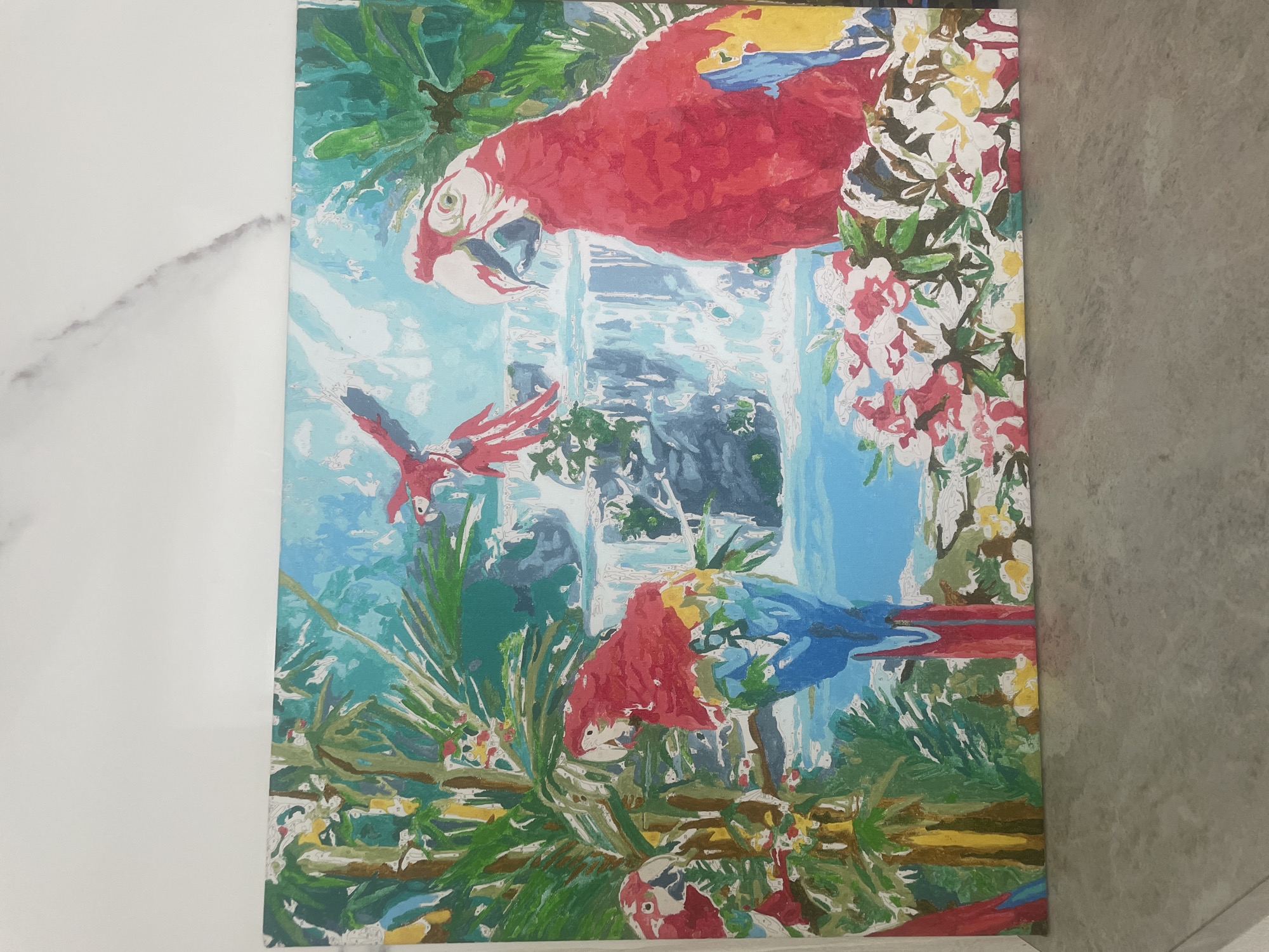The image is a painting of a tropical scene featuring parrots and a waterfall. The foreground contains lush green foliage and branches, with three parrots perched among them. One parrot is red and blue, facing slightly away from the viewer. Another parrot, mostly red, is lower in the composition and also perched on a branch. A third parrot is smaller and positioned near the bottom. Above the parrots, a red parrot is in flight. In the background, a waterfall cascades down, flanked by rocky cliffs and lush vegetation. The sky is a pale blue. The overall style of the painting is somewhat stylized and reminiscent of paint-by-number art. The painting appears to be resting against a wall, with a white marble surface visible on the left side and a gray stone surface on the right side. There is no indication of the city of Saratov, Russia.