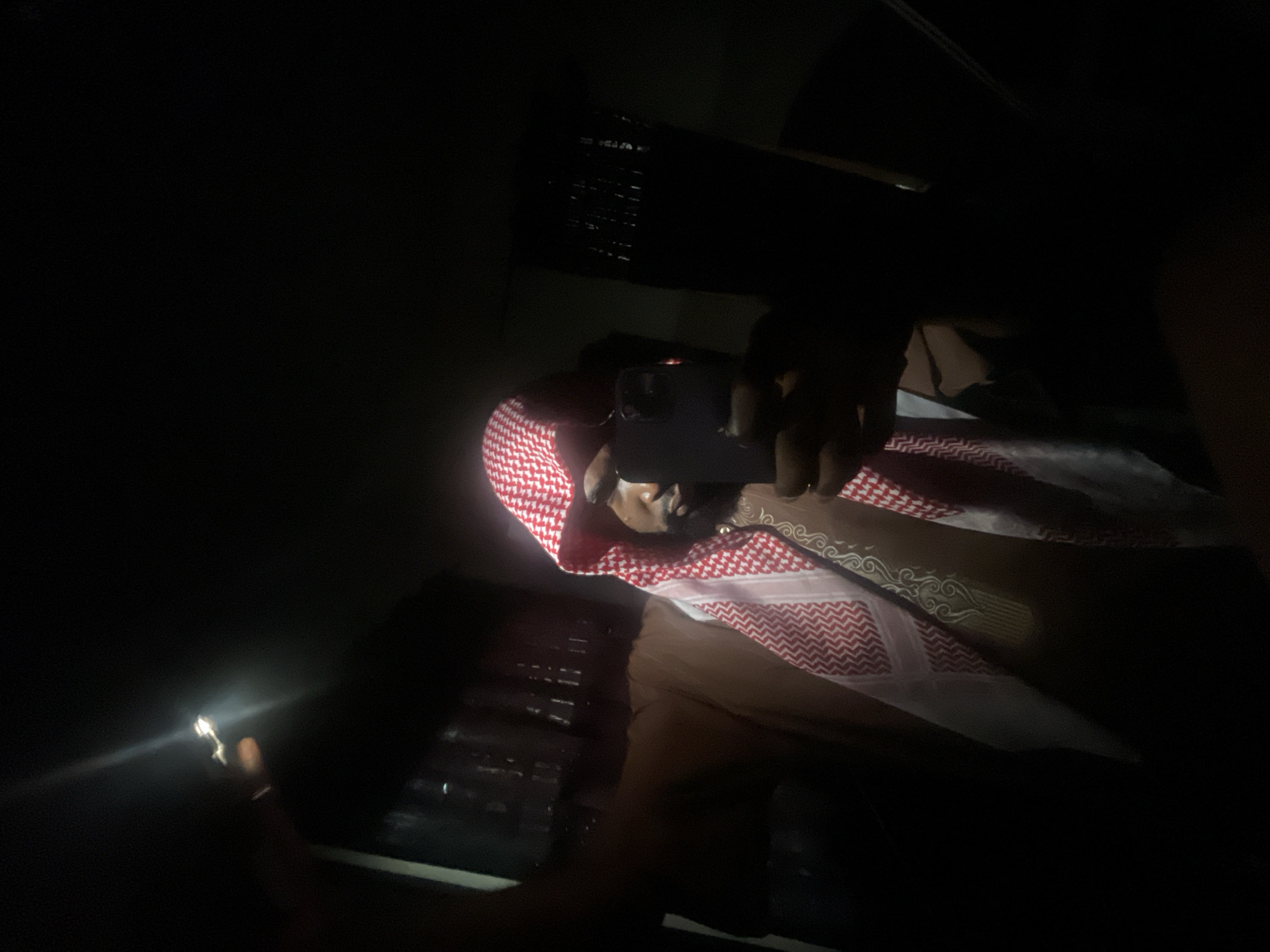 A person is shown at night in an indoor setting, likely a car. They are wearing a red and white checkered headscarf. The person is holding a smartphone and using the flashlight feature.