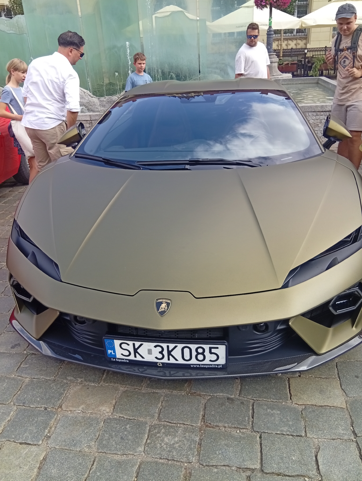 A matte gold Lamborghini is parked on a cobblestone street in Wrocław, Poland. The car's license plate reads "SK 3K085." A group of people are visible in the background, including a man in a white shirt, a child in a blue shirt, and two men in casual clothing. The scene appears to be a public area, possibly a plaza or street.