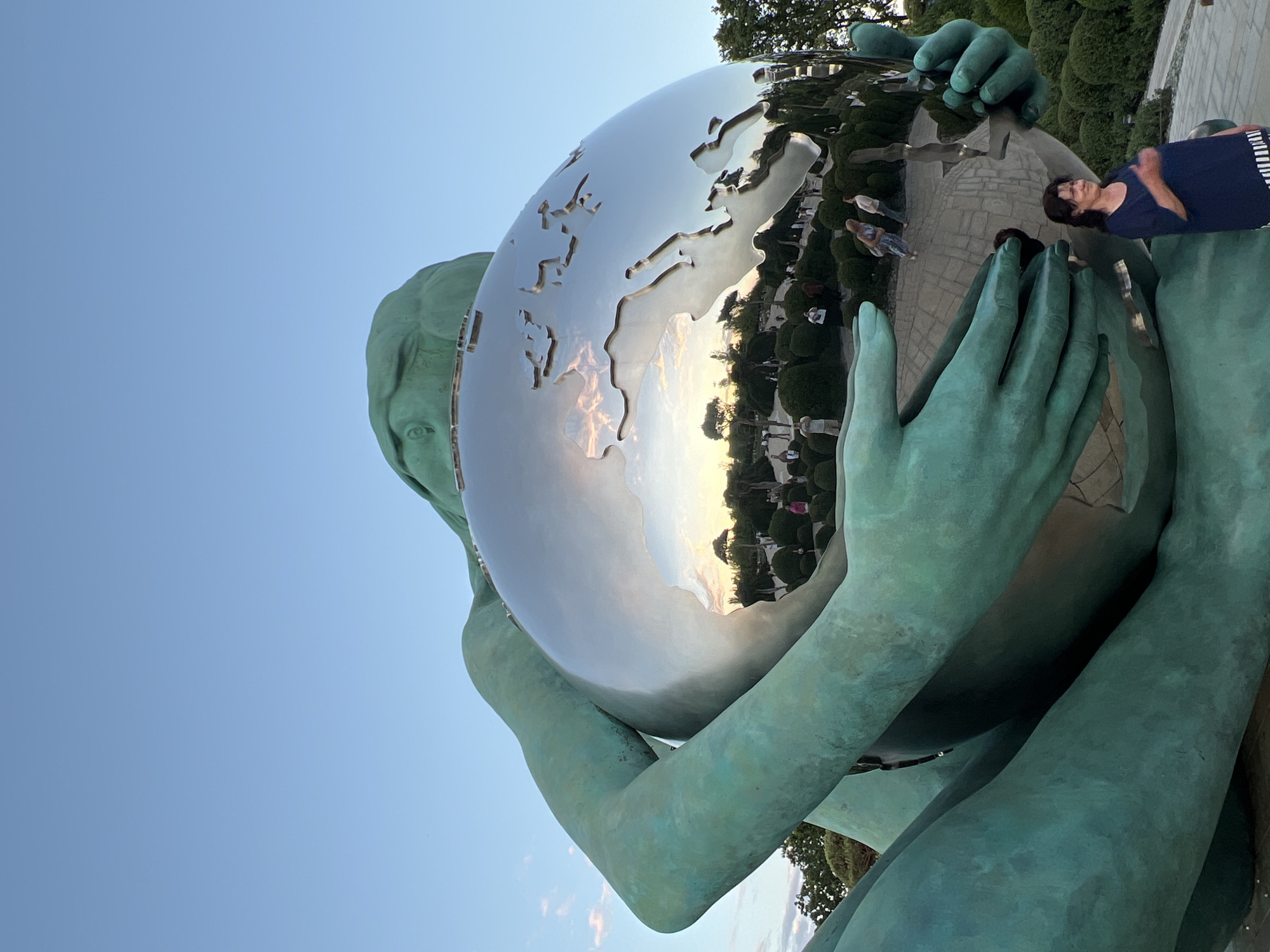 A large, turquoise statue depicting a stylized human figure cradling a mirrored globe is the central focus. The globe reflects the surrounding environment, including trees, bushes, a paved walkway, and several people. The sky above is clear and blue. In the lower right portion of the image, a woman in a dark blue shirt with striped trim stands looking towards the viewer, with her right hand raised. The statue is located in the city of Krasnodar, Russia.