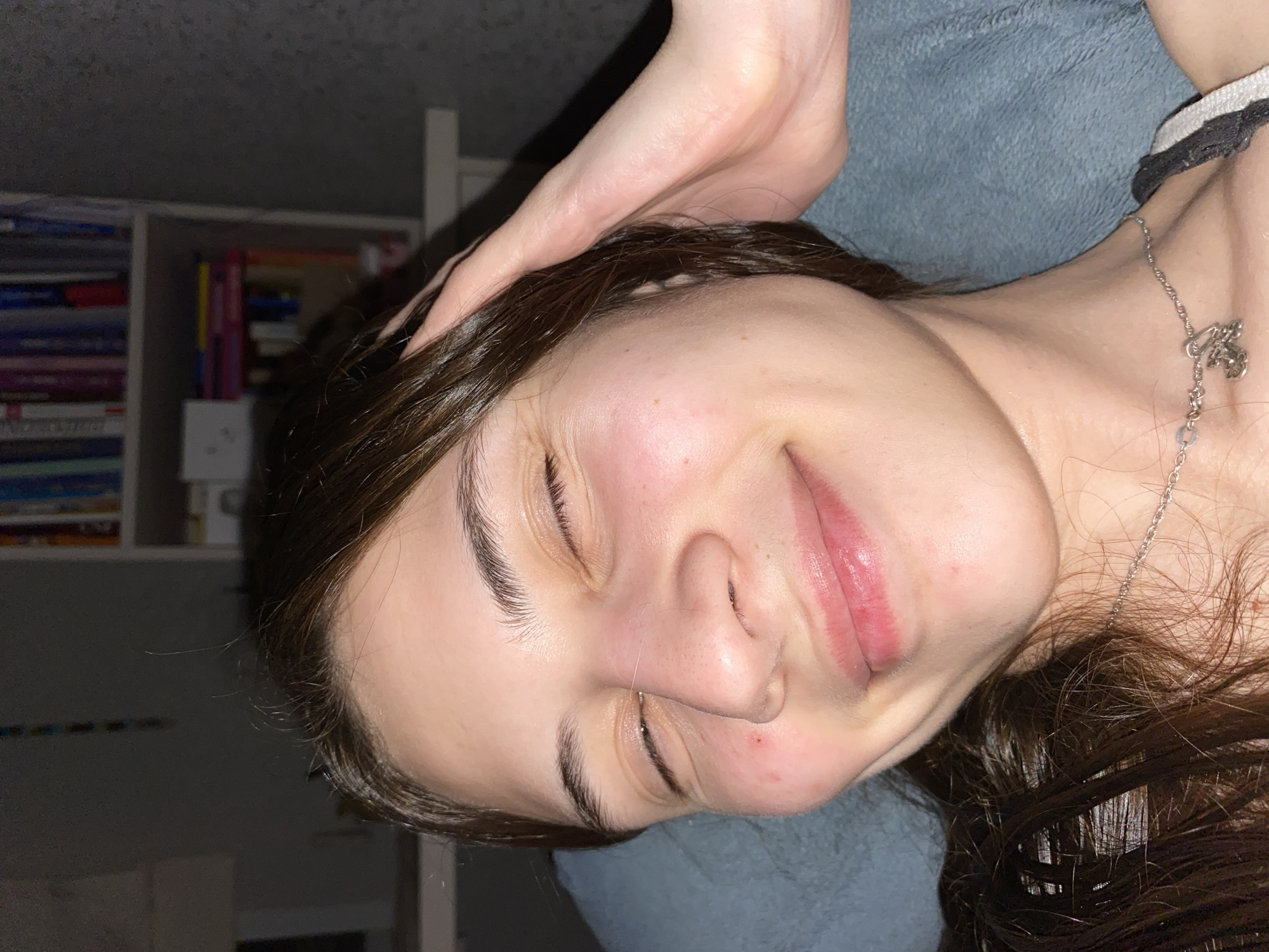 A young woman is lying down. Her head is turned to the right, and her eyes are closed. Her hair is brown. A hand is resting on the top of her head. She is wearing a silver necklace. The background includes a blue blanket, a bookshelf filled with books, and a white shelf. This image does not appear to be related to Votkinsk, Russia.