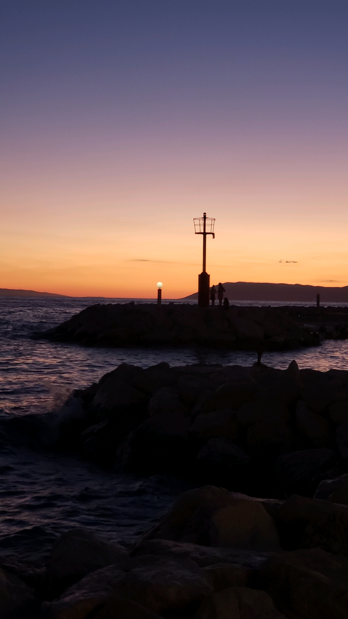 At dusk in Općina Tučepi, Croatia, a rocky pier extends into the sea. A tall, dark structure resembling a lighthouse or navigational marker stands silhouetted against the vibrant sunset sky. Three human figures and a dog are positioned on the pier near the base of the marker, appearing as dark silhouettes. A small, glowing orb is visible on a separate, shorter post further down the pier. The sea is active, with waves crashing against the rocks in the foreground. The sky transitions from a pale orange near the horizon to a deep purple overhead, indicating the end of daylight.