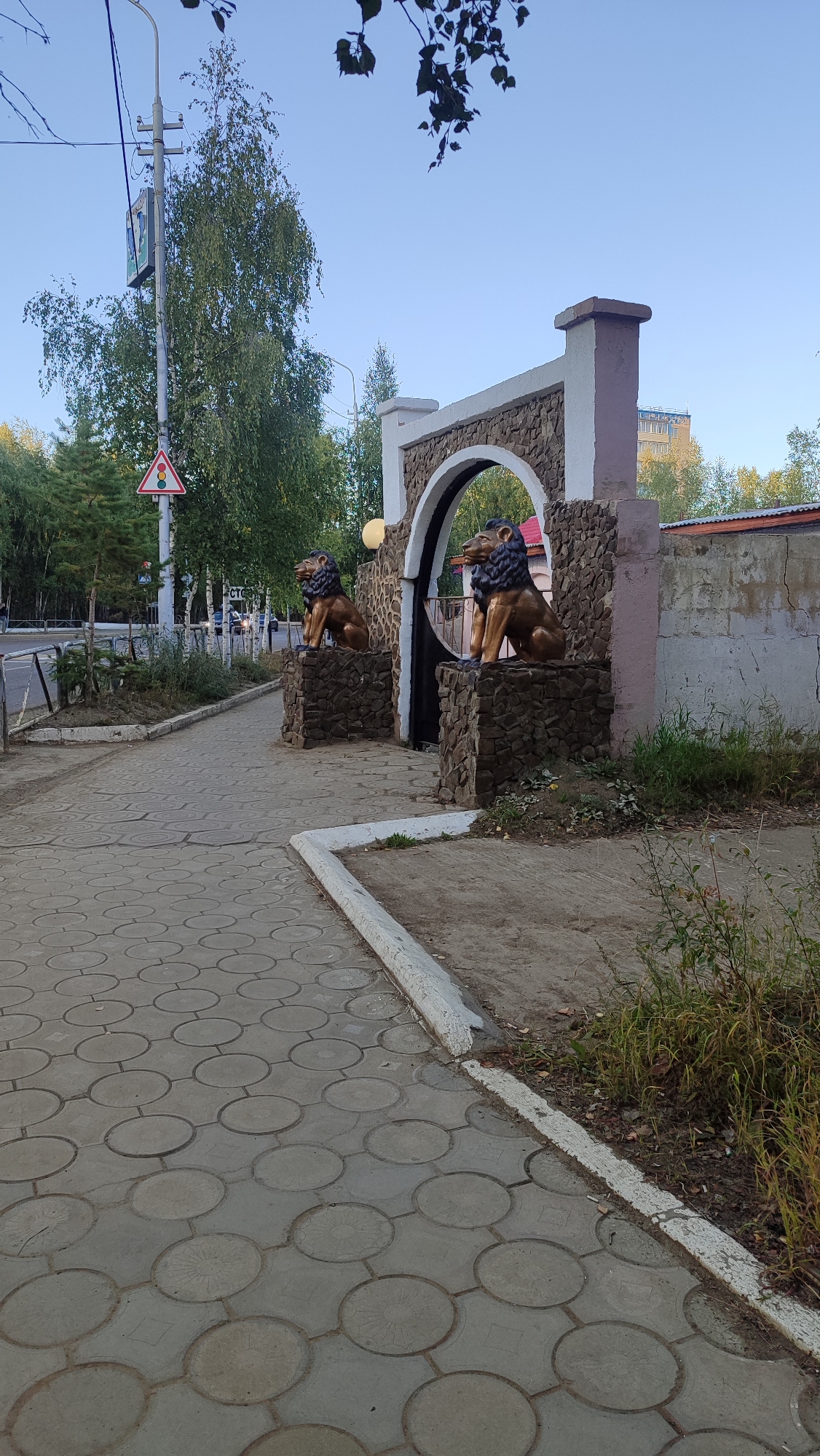 In Mirny, Russia, on a clear day, an arched gateway decorated with stone and flanked by two lion statues stands prominently. The gateway is situated on the right side of the image, leading into a dark entrance. The lion statues, made of a golden material with dark manes, are seated on stone pedestals. To the left of the gateway, a patterned concrete walkway leads away from the viewer. In the background, trees and a tall building are visible under a clear blue sky. A traffic light and a signpost are also present on the left side of the scene, near the road. The environment appears to be an urban setting with a mix of natural elements and man-made structures.