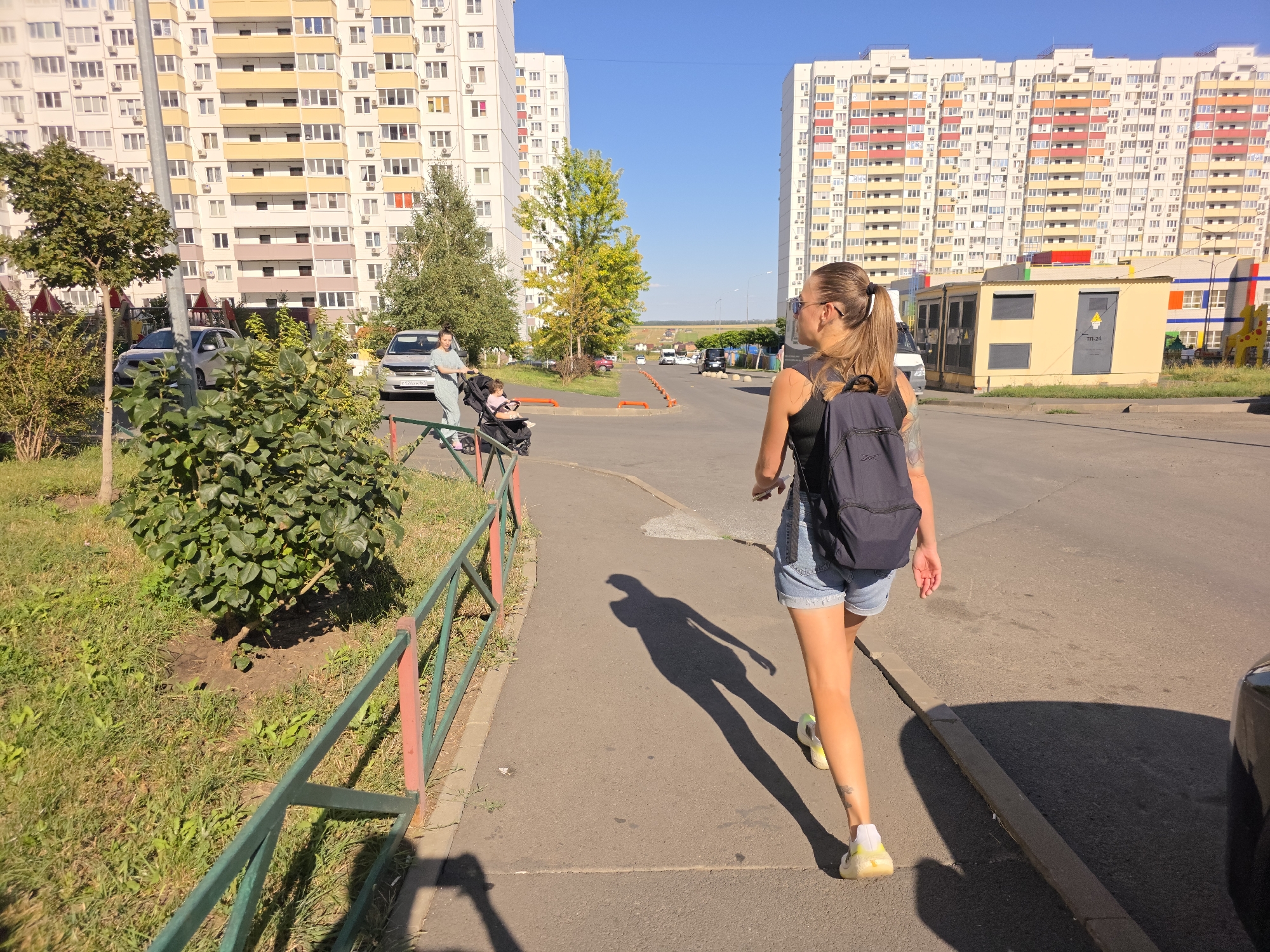 In Rostov-on-Don, Russia, a young woman with a backpack walks down a paved sidewalk next to a grassy area bordered by a green and red fence. She is wearing denim shorts and white sneakers. She has tattoos on her right arm. The sun casts a long shadow of the woman on the ground. Behind her, across the street, are two large apartment buildings, one yellow and white, and the other with colorful sections of red, orange, and yellow. There are cars parked on the street and a baby stroller visible on the sidewalk in the background. The sky is clear and blue.