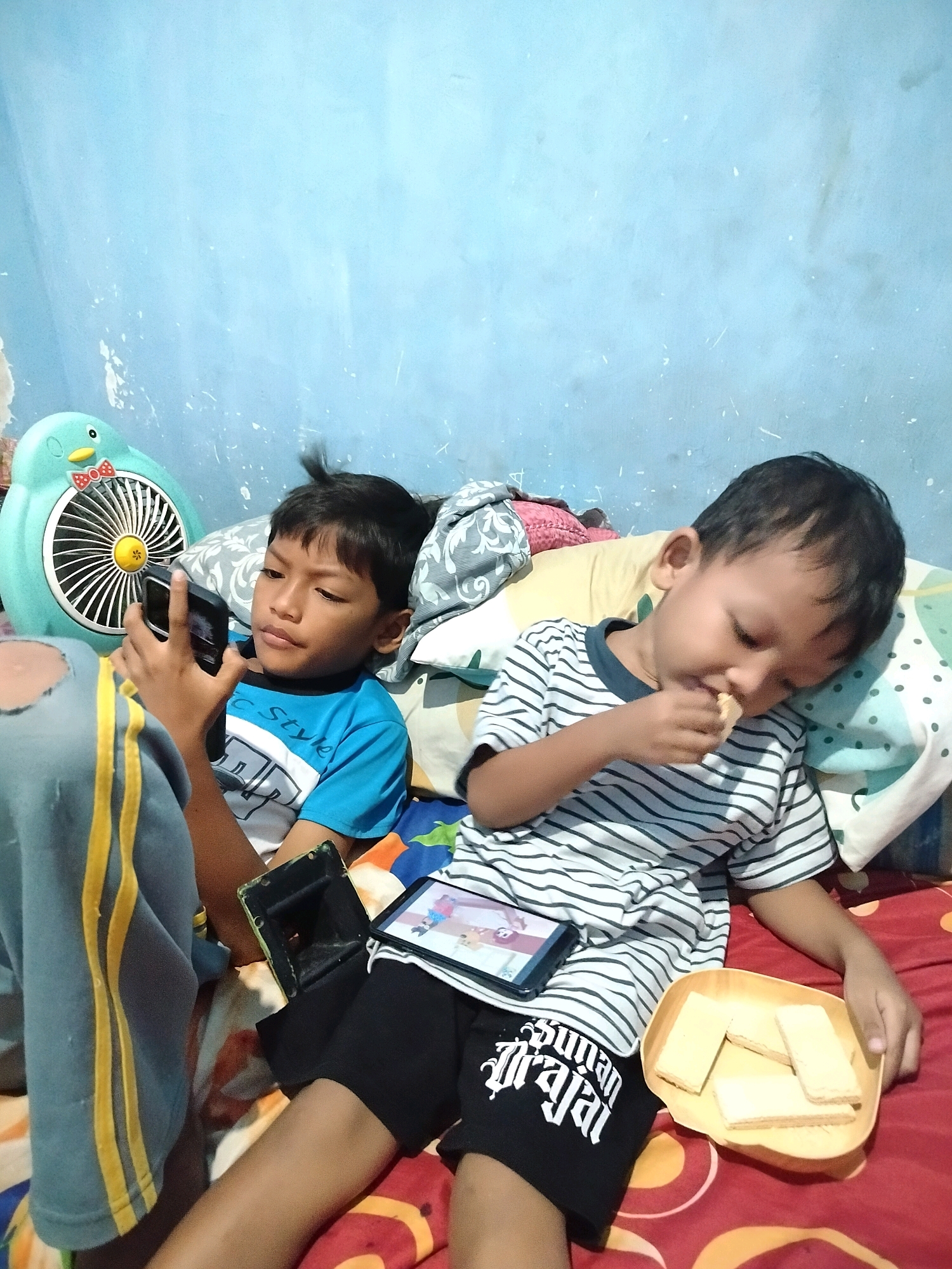 In a bedroom in Beloh, Indonesia, two young boys are on a bed. The boy on the left, wearing a blue and white t-shirt, is holding a smartphone. The boy on the right, in a striped t-shirt, is eating a wafer from a yellow dish. There is a blue penguin-shaped fan on the left side of the bed. The walls are a light blue color.