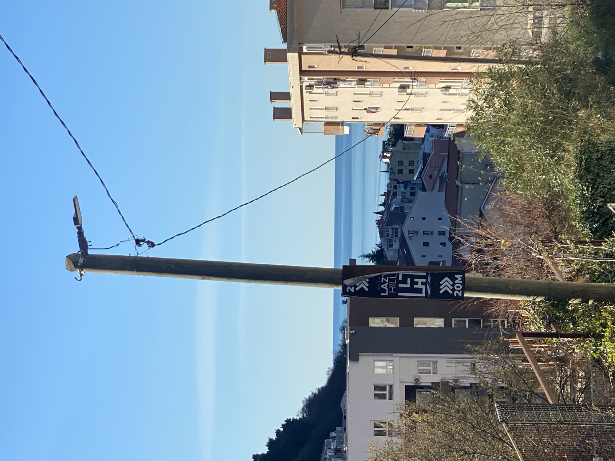 The image shows a slightly tilted outdoor scene in Budva, Montenegro. A wooden utility pole occupies the left side of the frame, topped with a streetlight fixture and strung with power lines that stretch across the top of the image. Attached to the pole is a sign indicating "Lazy Hill" with a direction arrow and "20M", suggesting a distance of 20 meters. In the background, buildings of varying heights and architectural styles are visible, with the Adriatic Sea and a light blue sky beyond them. Some greenery is present, with trees and shrubs interspersed among the buildings. The overall impression is a slightly elevated perspective of a neighborhood in Budva.