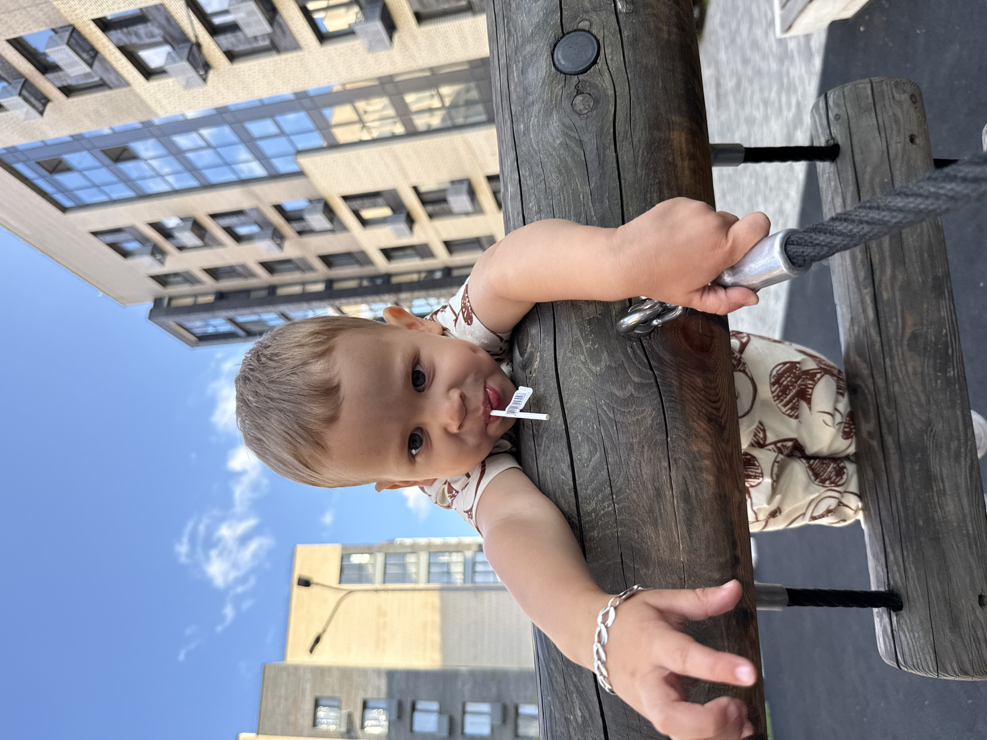 A young boy, likely between two and three years old, is shown at eye level, clinging to a wooden horizontal bar on a playground. He has blonde hair, dark eyes, and is holding a small white flag with a black and white sticker on a stick in his mouth. The boy is wearing a light-colored t-shirt with a pattern of brown shapes and matching light-colored shorts. A silver-colored bracelet is visible on his left wrist. His right hand is gripping a thick black rope attached to a metal fitting on the bar. In the background, modern, multi-story apartment buildings rise against a bright blue sky with scattered white clouds. The buildings have a light tan brick facade and numerous windows. The ground beneath the playground equipment appears to be dark, possibly asphalt or rubber surfacing. The overall scene suggests a sunny day in an urban playground setting, likely in Rusanovo, Russia, given the architectural style of the buildings and the presence of modern urban infrastructure.