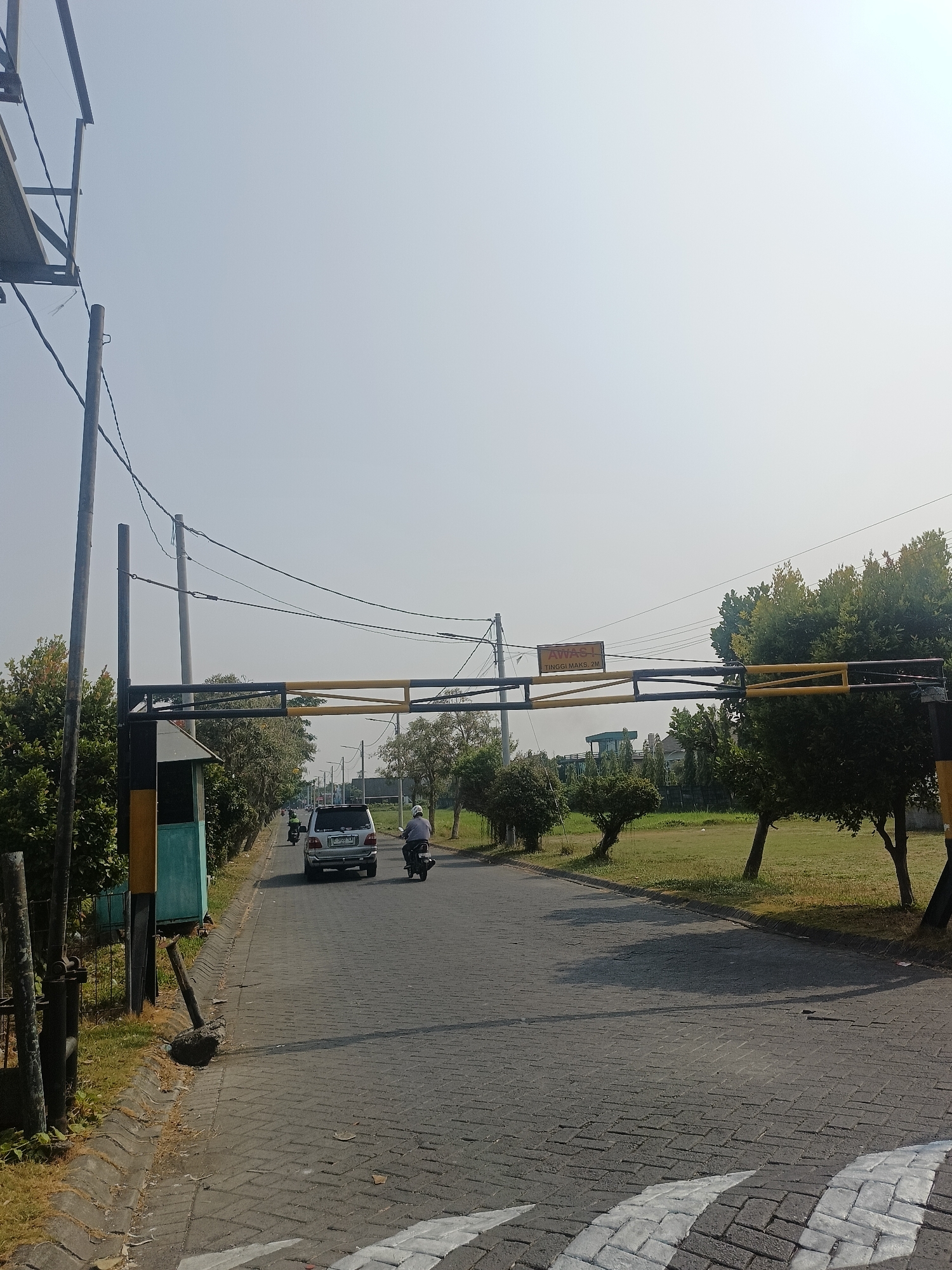 The image captures a street scene in Calukan RT 1 RW 5 Keboansikep, Indonesia, under a clear, hazy sky. A paved road, laid with interlocking brick pavers, forms the primary surface, with a section of white painted lines indicating a pedestrian crossing in the foreground. On the left side of the road, a small green and yellow booth with a metal frame stands adjacent to a grassy area and vegetation. A barrier, constructed with black and yellow horizontal bars supported by metal poles, stretches across the road, indicating a checkpoint or entrance. A sign with yellow lettering on a dark background is affixed to the barrier, displaying the text "AWAS-1 TINGGI MAKS, 2M," which translates to "CAUTION-1 MAX HEIGHT, 2M."
Traffic is present on the road. A silver SUV is visible in the middle distance, and a person on a motorcycle is proceeding in the same direction, slightly ahead of the SUV. Another motorcycle is further down the road. Electrical wires crisscross the sky, supported by tall poles on either side of the road. The environment includes lush green trees and grassy areas on the right side of the road, with some residential or commercial structures in the background. The overall impression is that of a moderately trafficked road leading into a developed area, possibly a residential complex or a controlled access point.