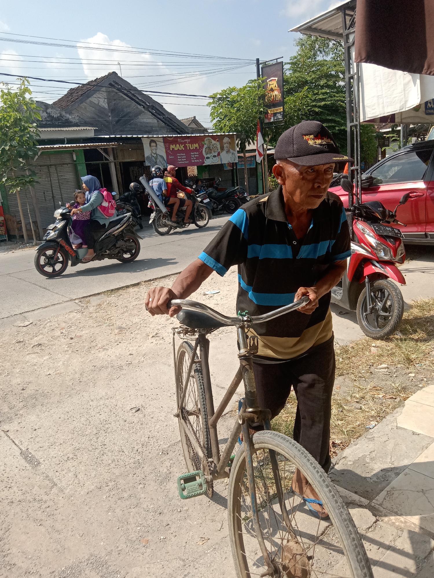 An elderly Indonesian man wearing a black baseball cap and a striped short-sleeved shirt with blue and black horizontal stripes and a tan panel over the shoulders stands next to an old bicycle on a dusty street in Klepek, Indonesia. The man is holding the handlebars of the bicycle with his right hand. He is wearing dark pants and blue flip-flops. In the background, a red car and several motorcycles are parked along the street. A woman and a child are riding a motorcycle on the left side of the frame. Buildings with corrugated metal roofs and banners are visible in the background. The sky is blue with some clouds.