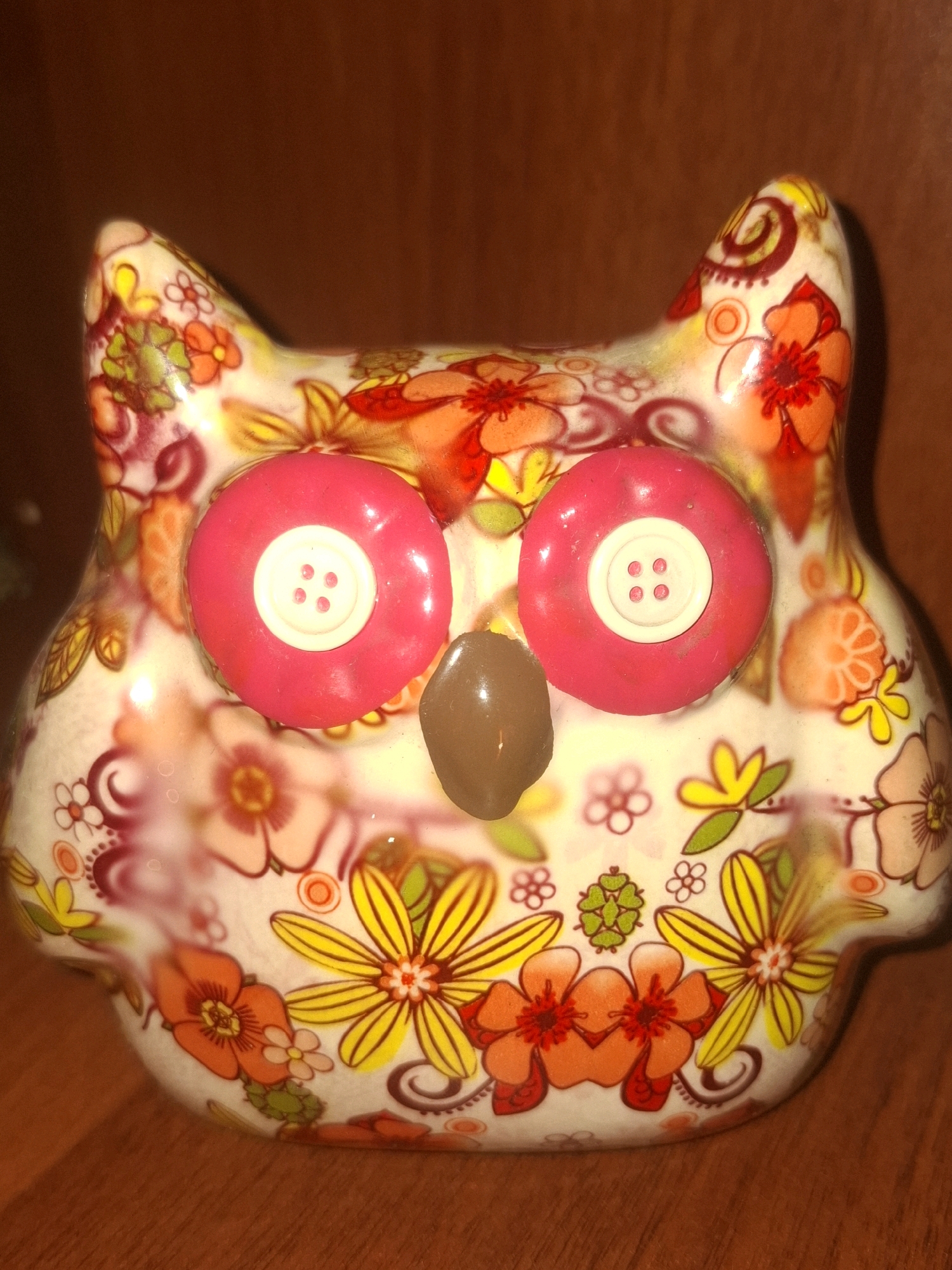 A ceramic owl figurine with a floral pattern is positioned on a wooden surface. The figurine has large, pink circular eyes with white button-like centers, and a brown, oval nose. The body of the owl is decorated with various flowers in red, orange, yellow, and pink hues, along with green leaves and swirling brown patterns. The background is a blurry, dark brown, wooden surface. This image does not depict a specific location in the city of Almaty, Kazakhstan.