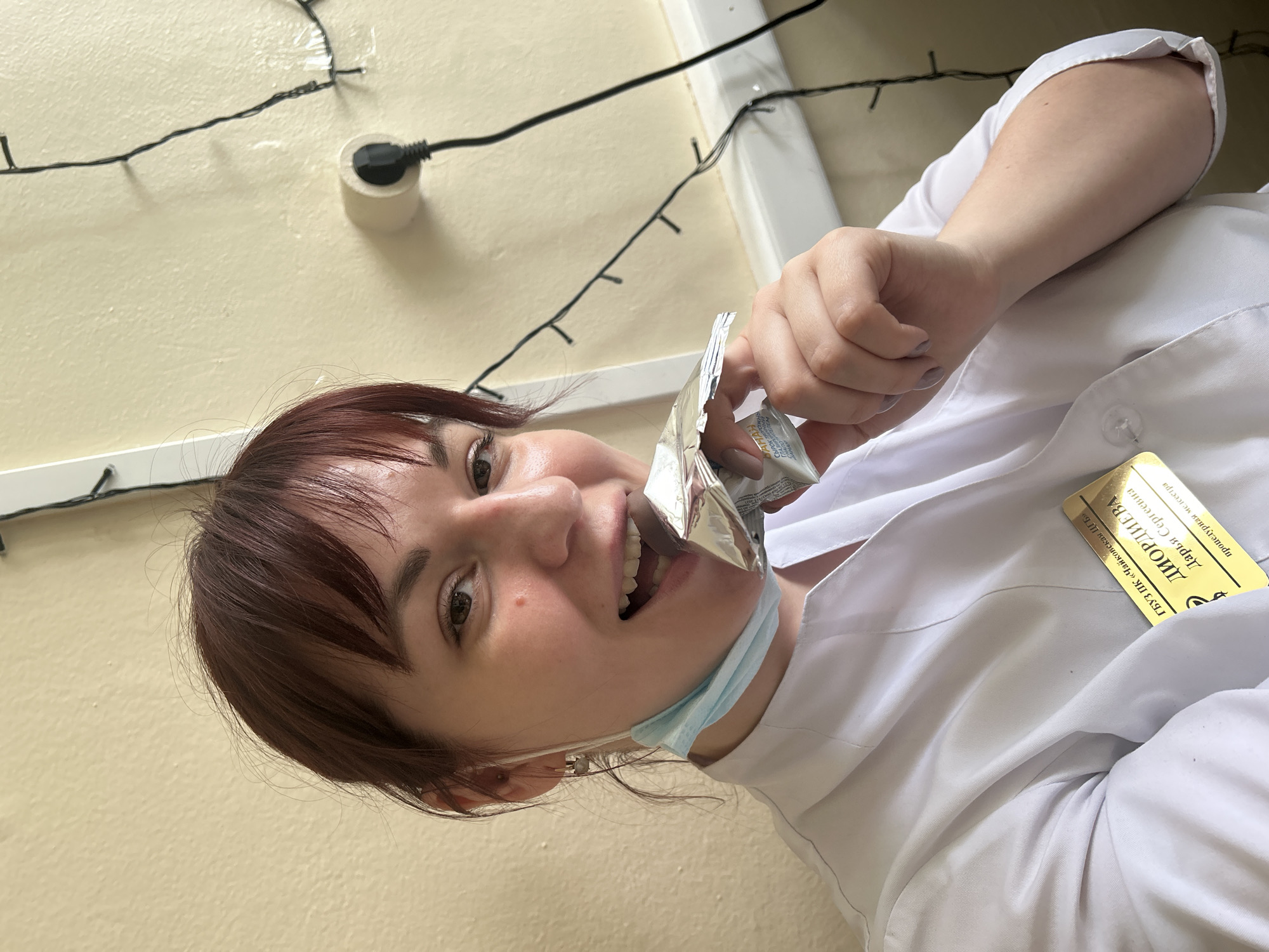 A woman, likely a medical professional, is seen from a low-angle perspective. She's wearing a white lab coat and a surgical mask pulled down below her chin. She's holding a partially unwrapped, foil-covered snack and appears to be about to take a bite. A name tag on her coat reads "ДИОРДИЕВА Дарья Сергеевна," indicating she is Дарья Сергеевна. The name tag also identifies her as a "процедурная медсестра" (procedural nurse) at the "ГБУЗ ПК «Чайковская ЦГБ»" (State Budgetary Healthcare Institution of the Perm Territory "Tchaikovsky Central City Hospital"). A portion of the snack package with the word Сырок is visible. A wall is in the background, with a string of lights near the ceiling. This could indicate the city of Чайковский, Russia, where the Tchaikovsky Central City Hospital is located. If the image was not taken at that hospital, the city is unknown.
