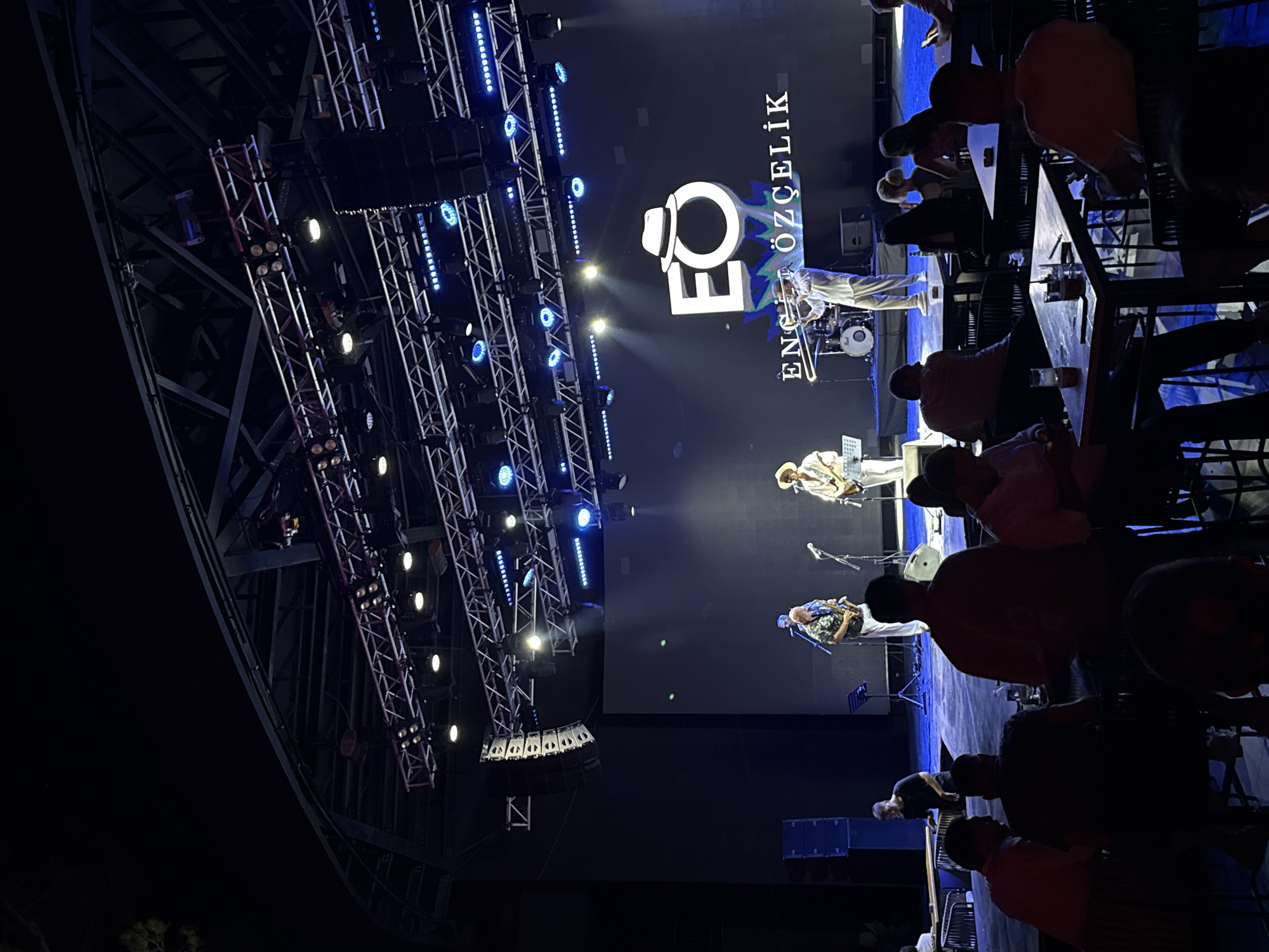 The image depicts an outdoor evening concert venue, likely in Bodrum, Turkey, given the presence of the "EO ENGE ÖZELIK" logo on the stage backdrop. The stage is illuminated with professional lighting, featuring a large screen displaying the artist's logo and name. A band of four musicians is performing on stage: a guitarist on the left, a saxophone player and a trombonist in the center, and another guitarist on the right. Several audience members are seated at tables in the foreground, enjoying drinks and observing the performance. The overall atmosphere is lively and engaging, suggesting a typical summer evening event.