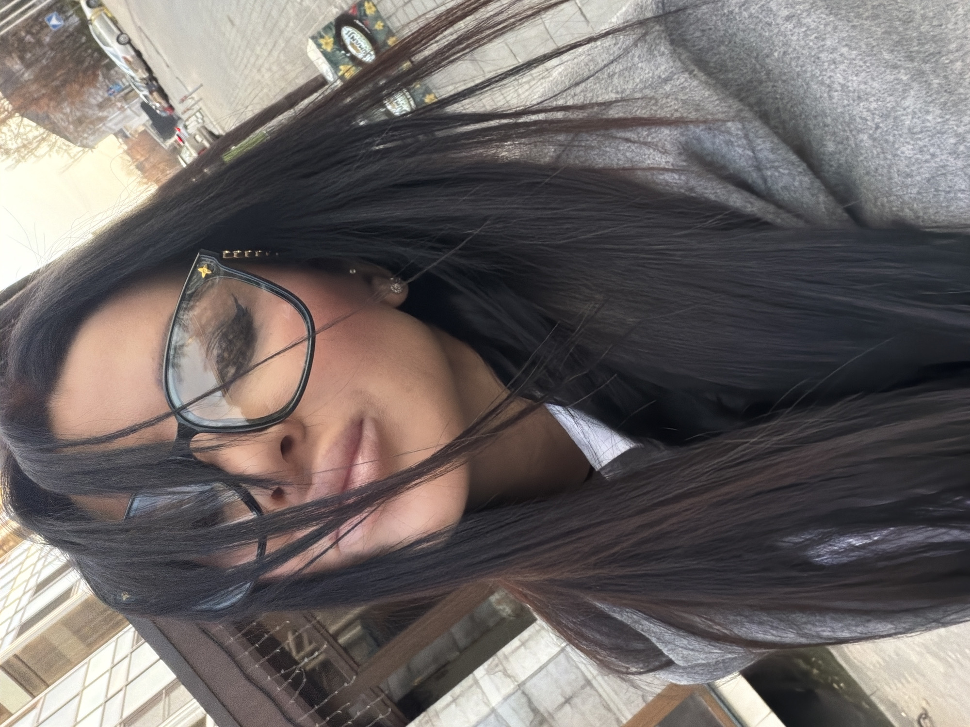 The image shows a person with long, dark hair and glasses. The person is wearing a gray garment. Outside the window or balcony, there is a view of a city street with vehicles and buildings. The location is Voronezh, Russia.