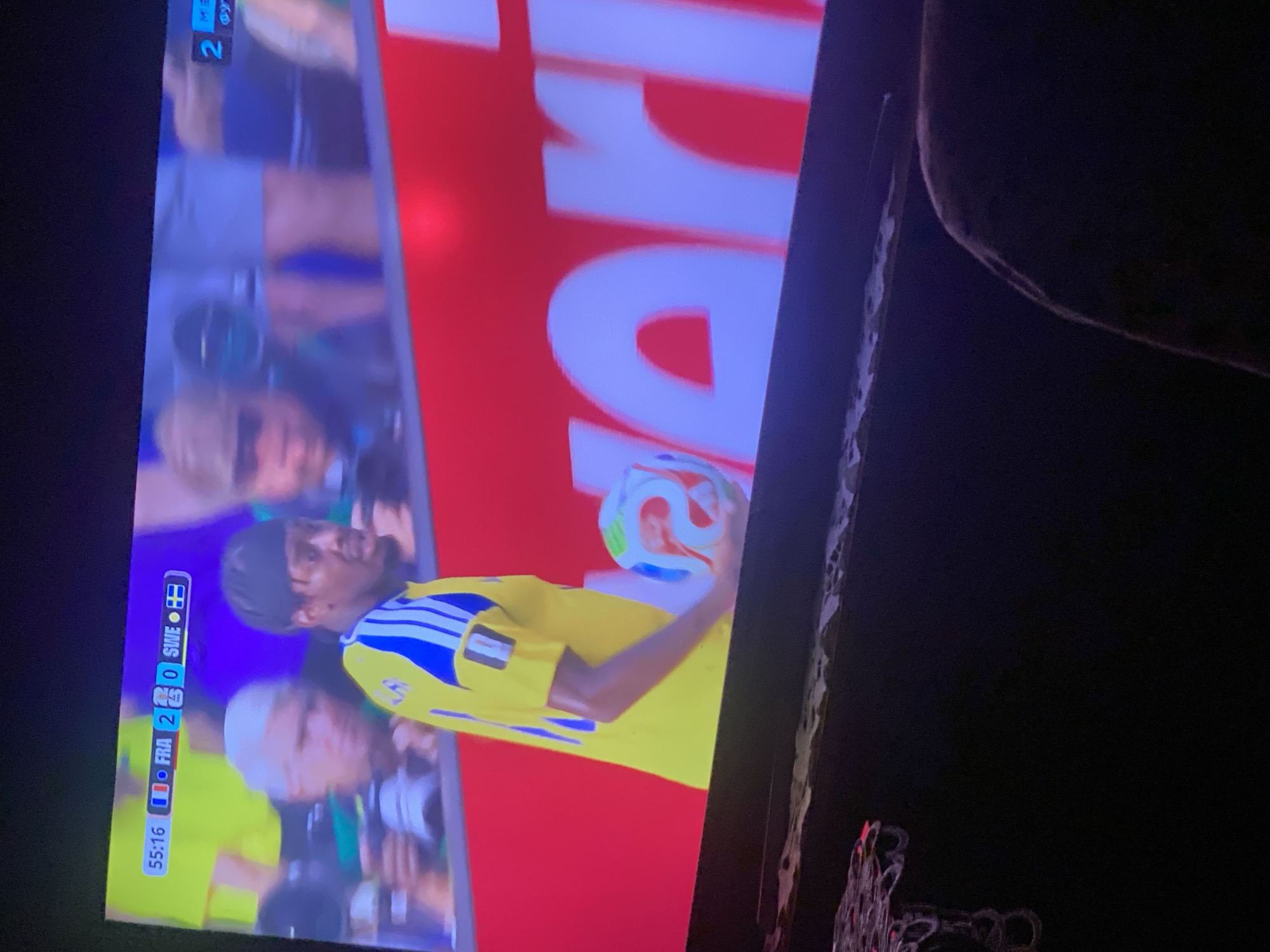 The image captures a sports match on a television screen. A football player in a yellow jersey with blue stripes is holding a multi-colored football. A scoreboard at the top of the screen shows "55:16" for the time, "FRA 2" for France, and "SWE 0" for Sweden, indicating the score and time of the game. The background shows blurred spectators and a red banner with white lettering. The television is placed on a dark surface, possibly a piece of furniture, and a person's leg is visible in the lower right corner, suggesting the image was taken from the perspective of an audience member. The scene is indoors, with the television screen being the primary focus. There is no indication of the location within the city of Zastavna, Ukraine.