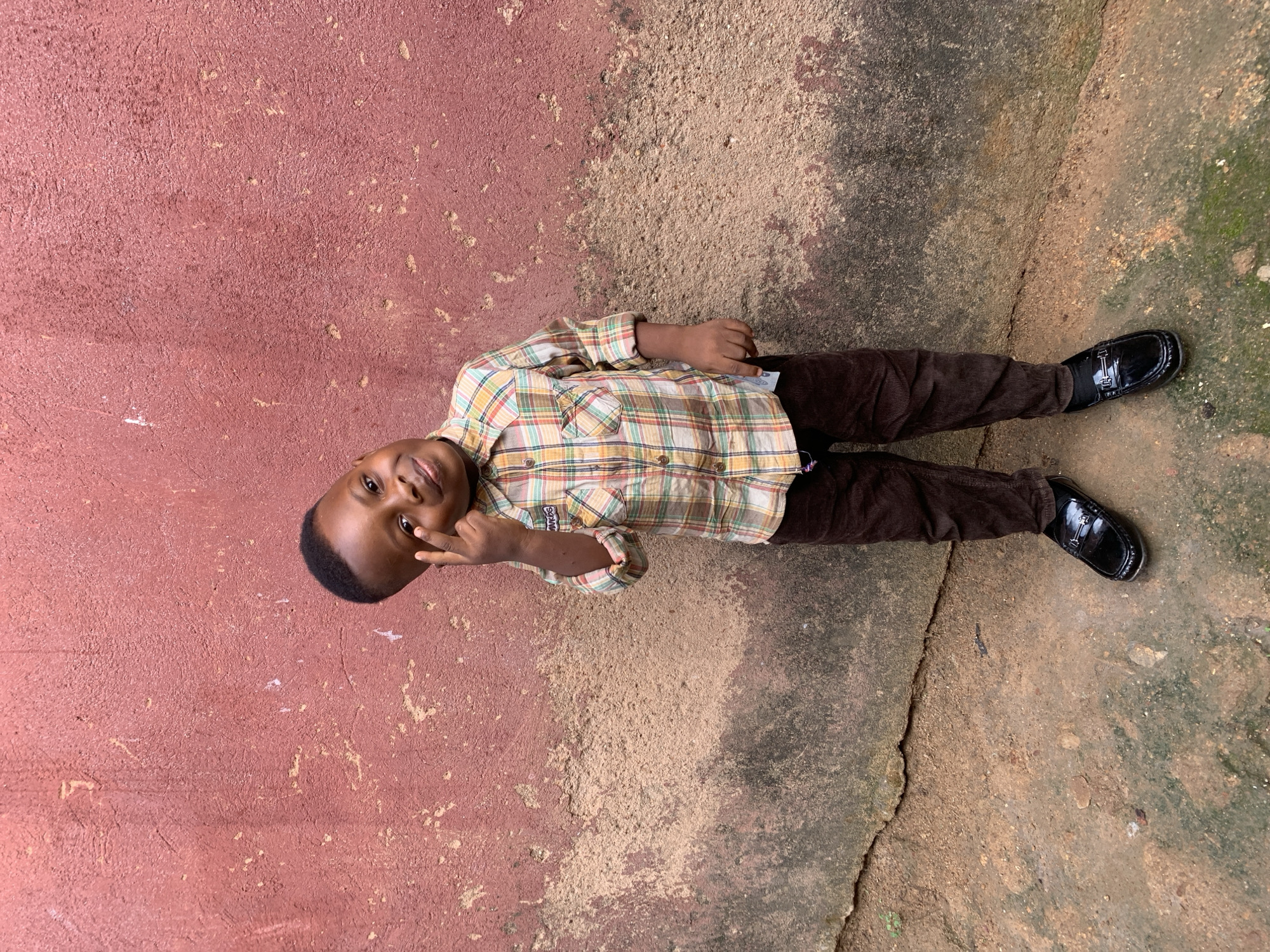 A young boy lies on the ground next to a textured red wall in Jos, Nigeria. He is wearing a plaid shirt, brown corduroy pants, and black dress shoes. The ground transitions from a light brown to a darker, weathered concrete with some patches of green. He is looking at the camera, holding up two fingers in a "peace" sign.