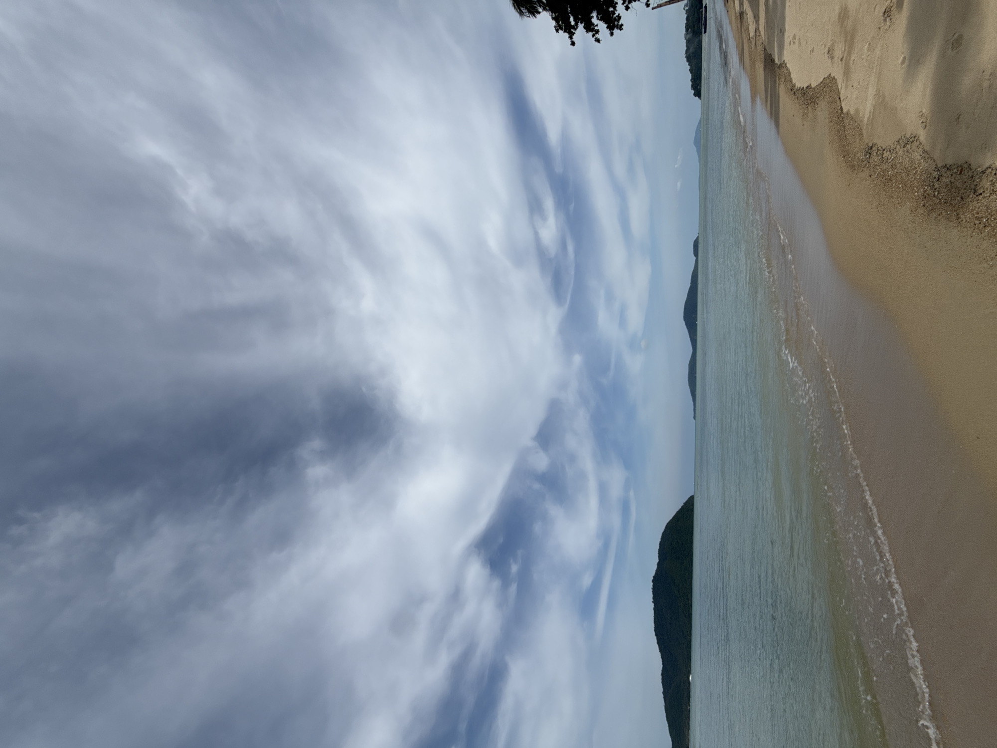 The image depicts a wide, sandy beach in Rawai, Thailand, with the calm, turquoise ocean lapping at the shore. The beach is mostly empty, with a few scattered footprints visible in the sand. In the distance, several lush green islands rise from the sea under a dramatic sky filled with swirling, wispy clouds. A dark silhouette of a tree is visible on the far right side of the frame, casting a shadow onto the sand. The overall scene is one of serene natural beauty.