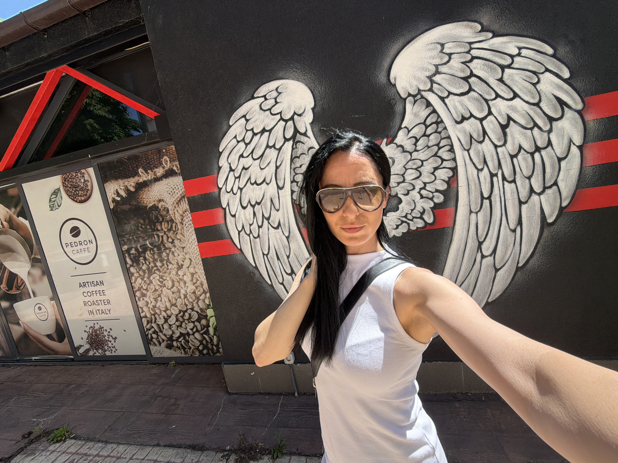 A woman is taking a selfie in front of a building with a mural. She is wearing a white tank top and aviator sunglasses. Her hair is black and pulled over to one side. Behind her, a mural on a black wall depicts a pair of large, stylized white angel wings, horizontally bisected by red stripes. To the left, a building façade displays advertising for "PEDRON CAFFE," describing it as "ARTISAN COFFEE ROASTER IN ITALY." The building has a black awning with red geometric accents. The ground is paved with dark gray bricks. The scene is outdoors, likely in the city of Pleven, Bulgaria, suggested by the Italian coffee advertisement.