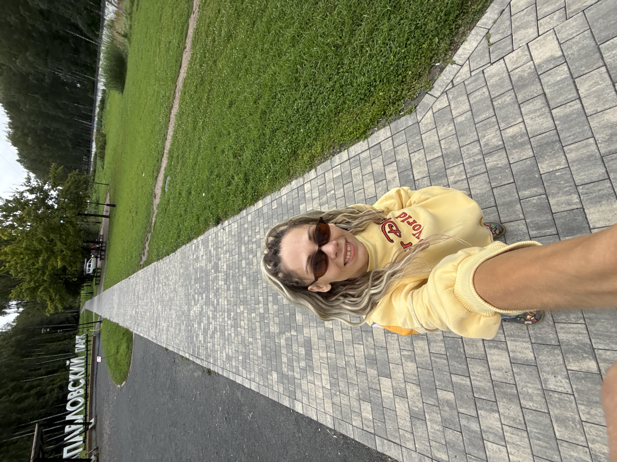 A woman with blonde hair and sunglasses is taking a selfie while standing on a paved walkway in Pavlovsk Park, Rusanovo, Russia. The walkway is made of light gray rectangular pavers. To the left of the walkway is a gravel area and a large white sign that reads "ПАВЛОВСКИЙ ПАРК" (Pavlovsk Park). Beyond the sign, a dense forest of birch trees lines a hill. To the right of the walkway is a lush green lawn and a body of water in the distance, with more trees and a cloudy sky overhead. The woman is wearing a yellow sweatshirt with red lettering. Her right arm is extended towards the camera, holding a selfie stick.