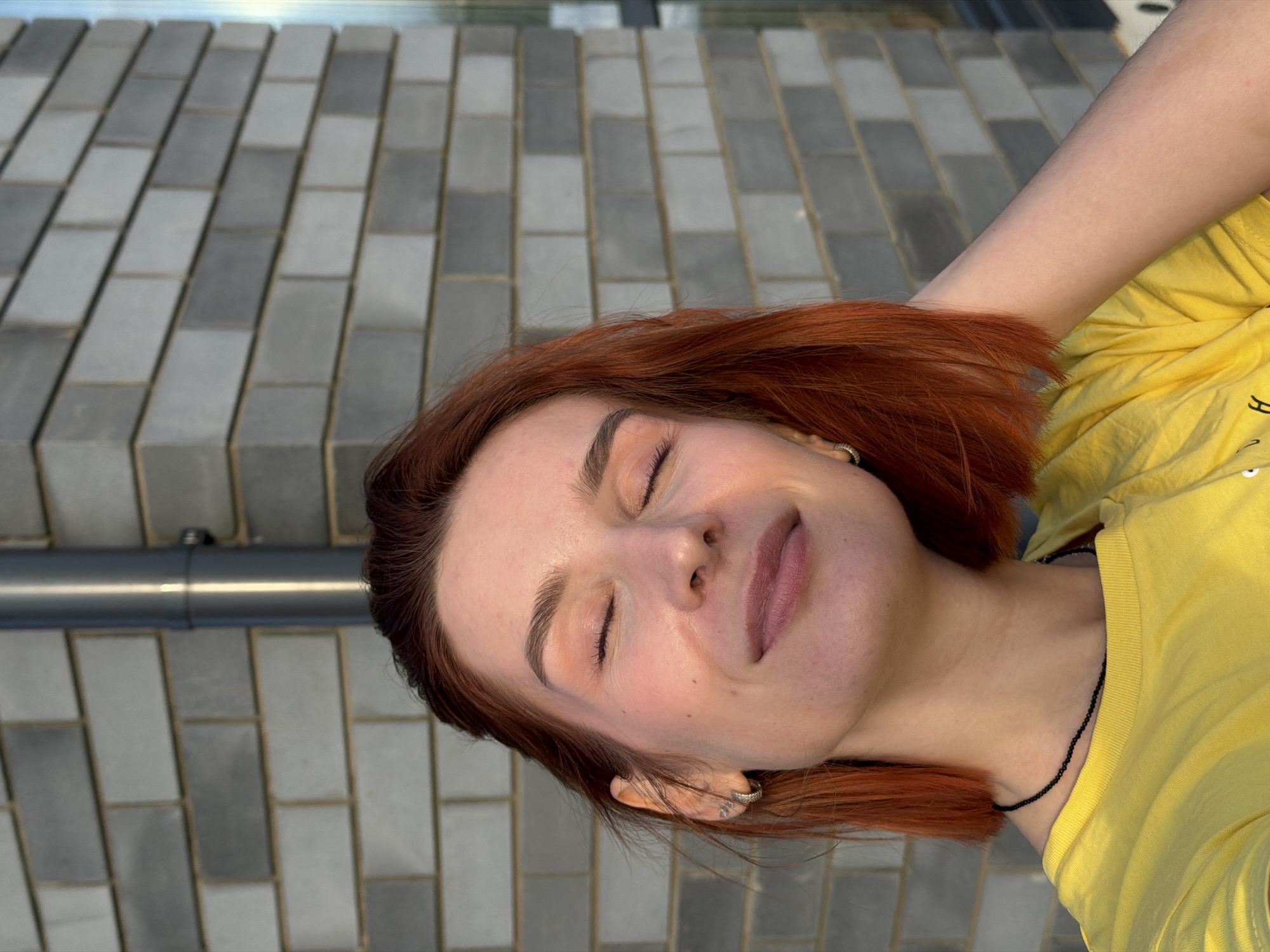 This image depicts an adult female positioned outdoors in Chaikovsky, Russia, captured from the chest up against a grey brick wall. She has short, auburn-colored hair and fair skin. Her eyes are closed, and she displays a subtle smile, with her head tilted slightly upward and to the left. The visible left ear is adorned with a small hoop earring and two stud earrings. She wears a yellow short-sleeved t-shirt and a thin, dark beaded necklace. Her right hand is partially visible, appearing to rest behind her neck. The background consists of a grey brick wall, constructed in a stretcher bond pattern, with a vertical grey metallic downspout affixed to its right side. A narrow, vertical reflective glass panel is partially visible to the far right. The scene is illuminated by natural light, casting a warm tone on the subject's face, indicating a daytime setting.