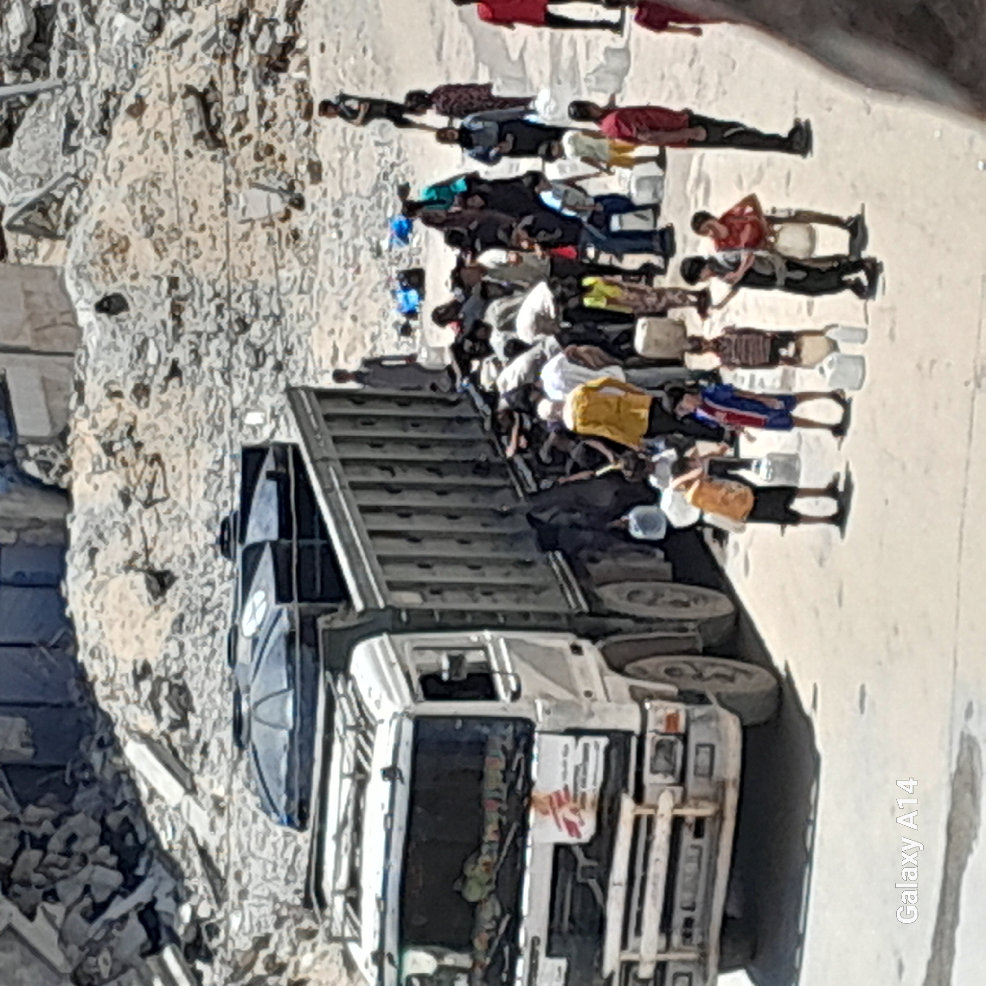 In the city of Khan Yunis, Palestinian Territories, a truck is parked near piles of rubble and damaged buildings. The truck is loaded with two large black water tanks and is surrounded by a group of people, many of whom are holding white plastic water containers. The people appear to be in the process of collecting water, with some individuals, including children, actively engaged in carrying or filling containers. The scene suggests a humanitarian effort to distribute water amidst destruction.