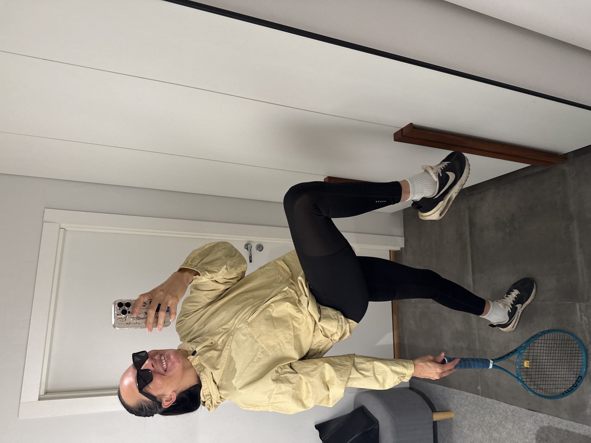 A person is standing in a room, taking a selfie in a mirror. They are wearing black sunglasses, a light yellow hooded jacket, black leggings, white socks, and black and white athletic shoes. They are holding a blue tennis racket in their left hand and their right hand is holding up a smartphone. The room has tiled flooring and white walls. There is a gray stool with wooden legs on the left side of the image and a tall white cabinet or closet on the right. The person's pose suggests they are in mid-action, perhaps jumping or about to jump. There is no indication of the city of Chaikovsky, Russia, in the image.