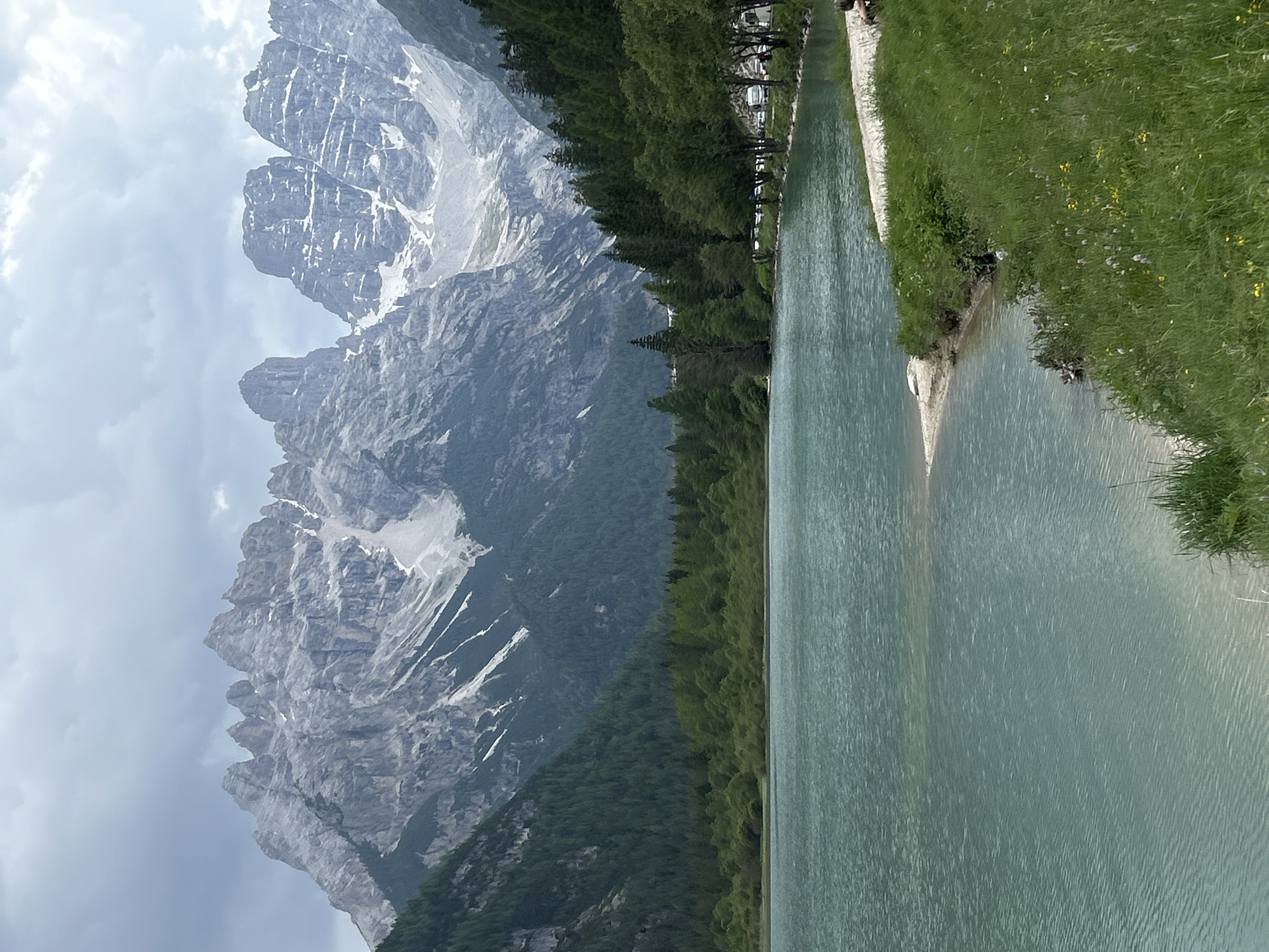 An outdoor, full shot captures a tranquil lake with a backdrop of dramatic, rocky mountains under a cloudy sky. The lake, a striking turquoise, fills the lower half of the frame, its surface rippled by a gentle breeze. A sandy shore curves along the right side, giving way to a grassy embankment dotted with small yellow and purple wildflowers. A paved walkway runs along the edge of the grass, where several cars are parked. In the far distance, the jagged peaks of the mountains, partially covered in snow, rise majestically, their grey rock faces contrasting with the lush green of the pine forests that climb their lower slopes. The sky is overcast with thick, grey clouds, creating a muted, atmospheric light.