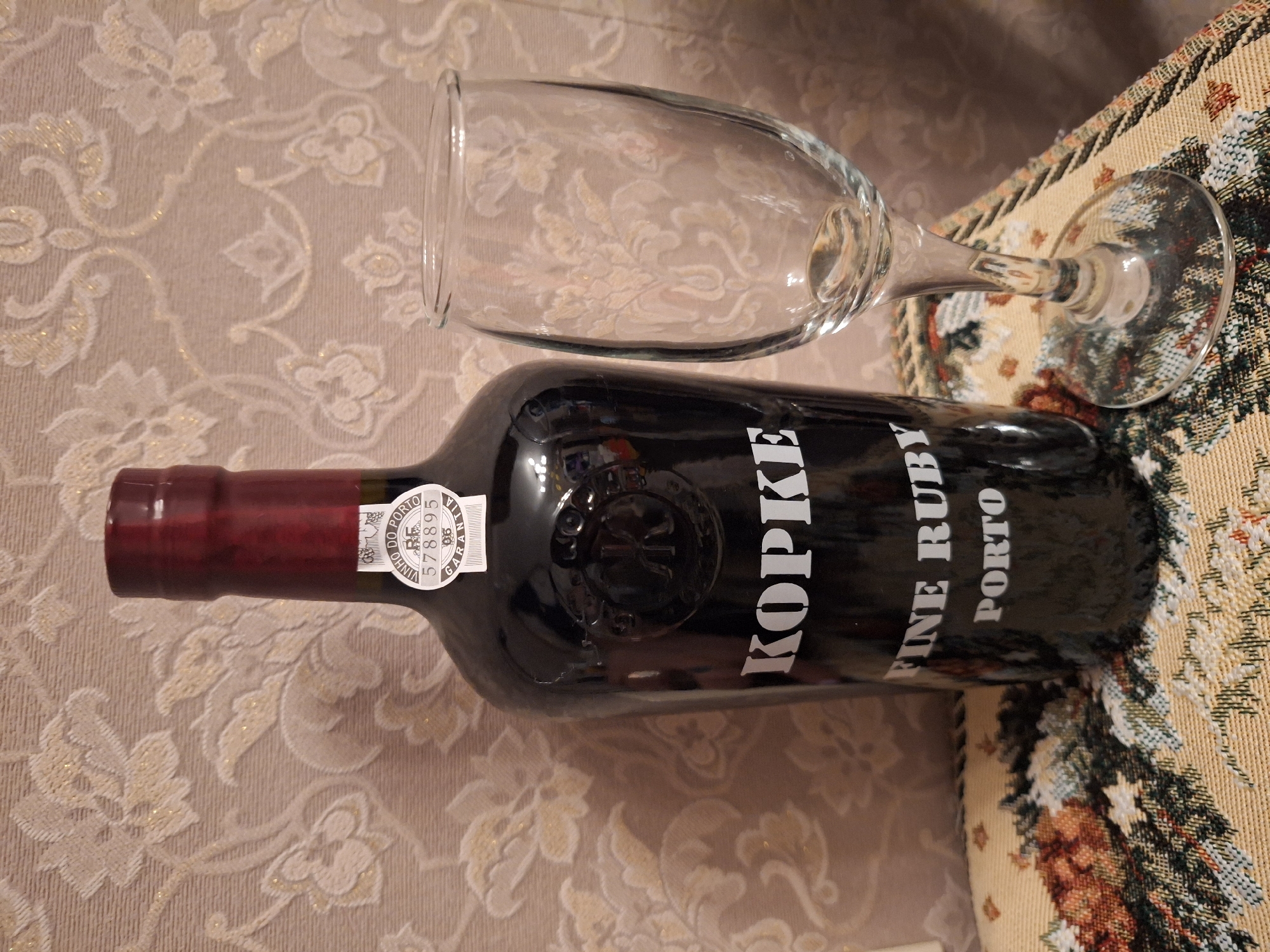 The image displays a dark-colored wine bottle and an empty, clear wine glass laid horizontally on a patterned surface. The bottle is prominent, featuring a main label with the brand "KOPKE" in large white capital letters, and "FINE RUBY PORTO" below it. An embossed logo or crest is also visible on the bottle's shoulder. On the bottle's neck, a white seal is present with text including "VINHO DO PORTO," a serial number "578895," and "GARANTIA," indicating its origin as Portuguese Port wine. The wine glass is simple and clean, suggesting readiness for use or recent use.
The objects are resting on two distinct textile surfaces. The primary background is a light, possibly beige or cream, material with a subtle, shimmering floral or damask-like pattern. To the right, the items overlap with a more densely woven or embroidered fabric, possibly a cushion or tapestry, which exhibits a colorful pattern of greens, browns, and yellows, resembling foliage or a landscape.
There are no people visible, and no active event is taking place, only the static arrangement of the bottle and glass. The setting appears to be an indoor environment. There are no visual cues within the image itself to confirm the provided location context of Moscow, Russia, beyond it being a domestic or indoor scene. The time of day and weather are not discernible.