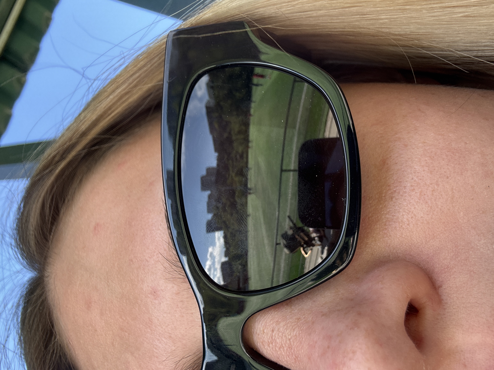Close-up, canted portrait of a blonde woman wearing black sunglasses. The reflection in the left lens reveals a cityscape and a green space. The right lens shows a blurry object. The woman has fair skin. The sky is visible in the upper-left corner. Considering the architectural style reflected in the sunglasses, the city is likely Tbilisi, Georgia, specifically near a park or open green area that offers views of the city's skyline.