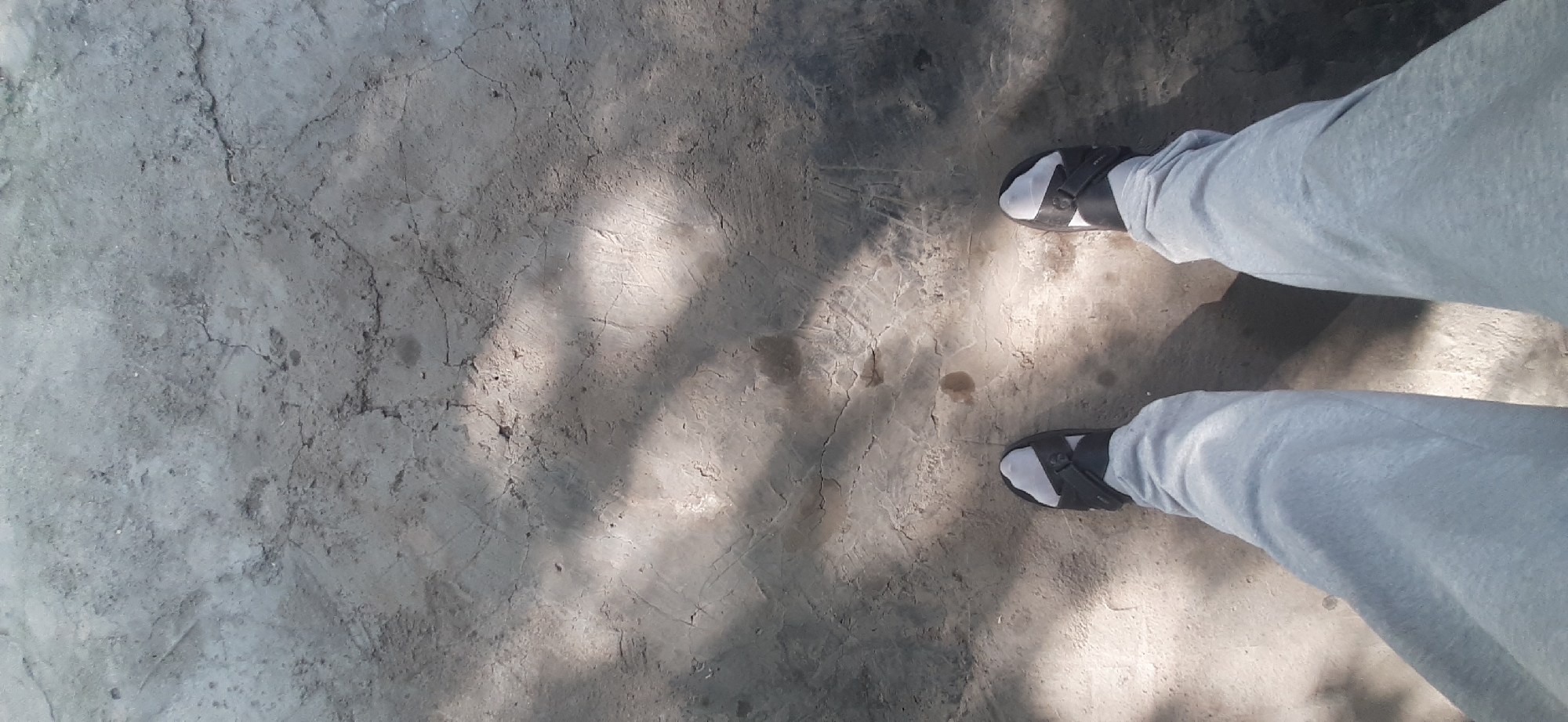 A low-angle, overhead shot captures a person's legs and feet standing on a cracked, concrete surface. The concrete is predominantly gray with visible lines of fracture and some darker, wet-looking patches. Sunlight filters through what appears to be foliage, casting dappled shadows across the ground. The individual is wearing light gray sweatpants and black slide sandals with white socks. The scene is static, with no discernible action taking place. The environment suggests an outdoor, urban or semi-urban setting, possibly a walkway or public area. The image does not provide specific Yessentukskaya, Russia, landmarks or features.