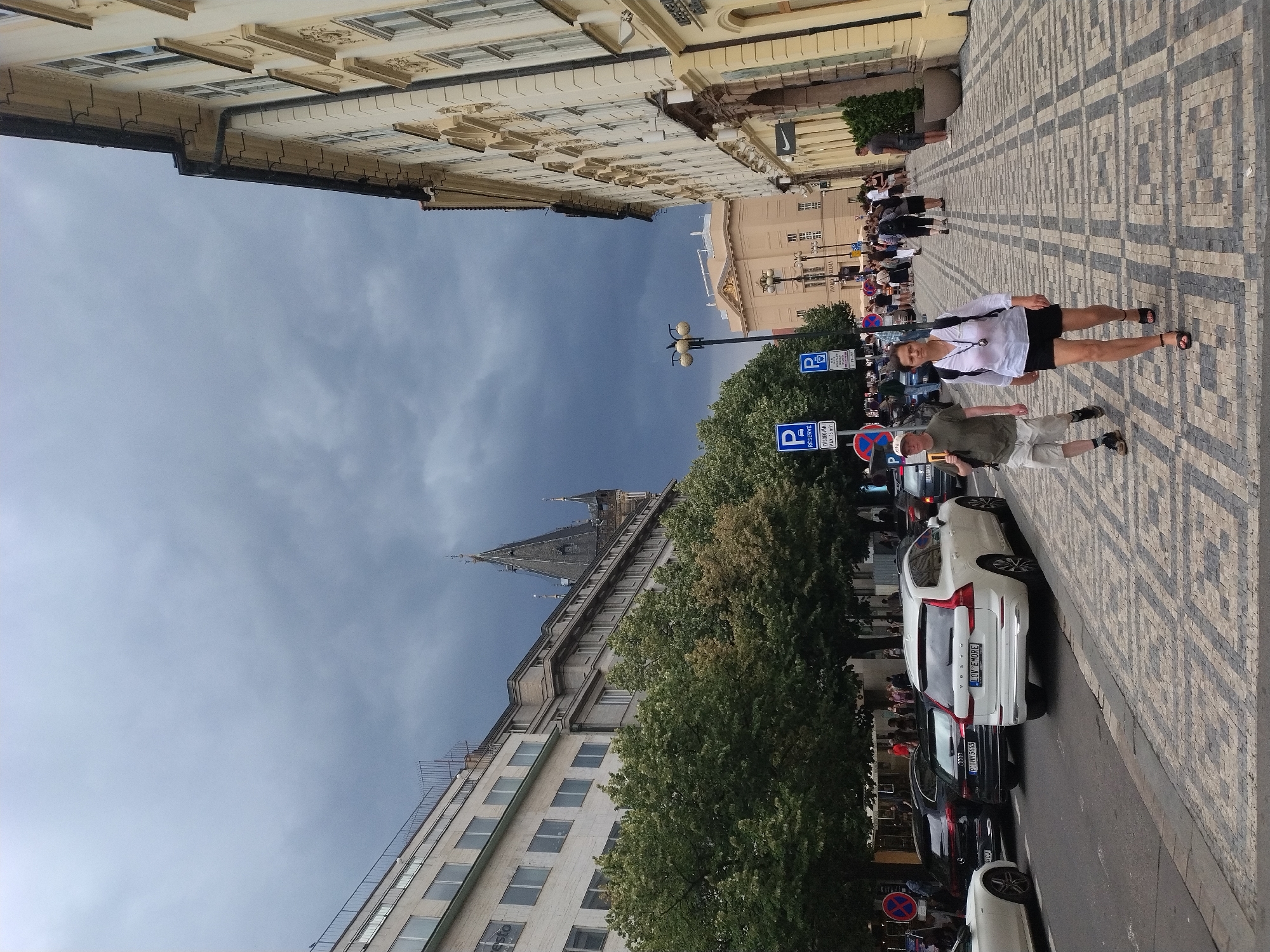 This is a medium-shot, eye-level, outdoor photograph taken on a city street, likely in Prague, Czechia, judging by the architecture and street signage. The scene depicts a cobblestone sidewalk with a paved street running alongside. In the foreground, two individuals are walking towards the viewer on the sidewalk. The person on the left is a man wearing a light-colored t-shirt, beige shorts, a baseball cap, and dark shoes. He has a backpack on his left shoulder. The person on the right is a woman wearing a white short-sleeved blouse, a black skirt, and black heeled sandals. She is also carrying a backpack on her right shoulder.
Beyond the individuals, the sidewalk is lined with trees that have full green foliage. Further down the street, several cars are parked. A white Volvo SUV with a license plate reading "LOV EMOFE" is prominent in the mid-ground on the right. Other vehicles, including a black Audi SUV and a white sedan, are visible on the left side of the street.
The background features a series of buildings with a mix of architectural styles. On the left, a modern-looking building with multiple windows and a balcony-like structure is visible. Adjacent to it, a more ornate building with a dark, pointed spire rises into the sky. On the right side of the street, a pale yellow building with decorative elements and numerous windows lines the sidewalk. Further in the distance, another light-colored building with a prominent facade and columns can be seen. The sky is overcast with dark, dramatic clouds, suggesting an impending storm. Streetlights and parking signs are also present along the street. The overall atmosphere is that of a busy urban environment under a brooding sky.