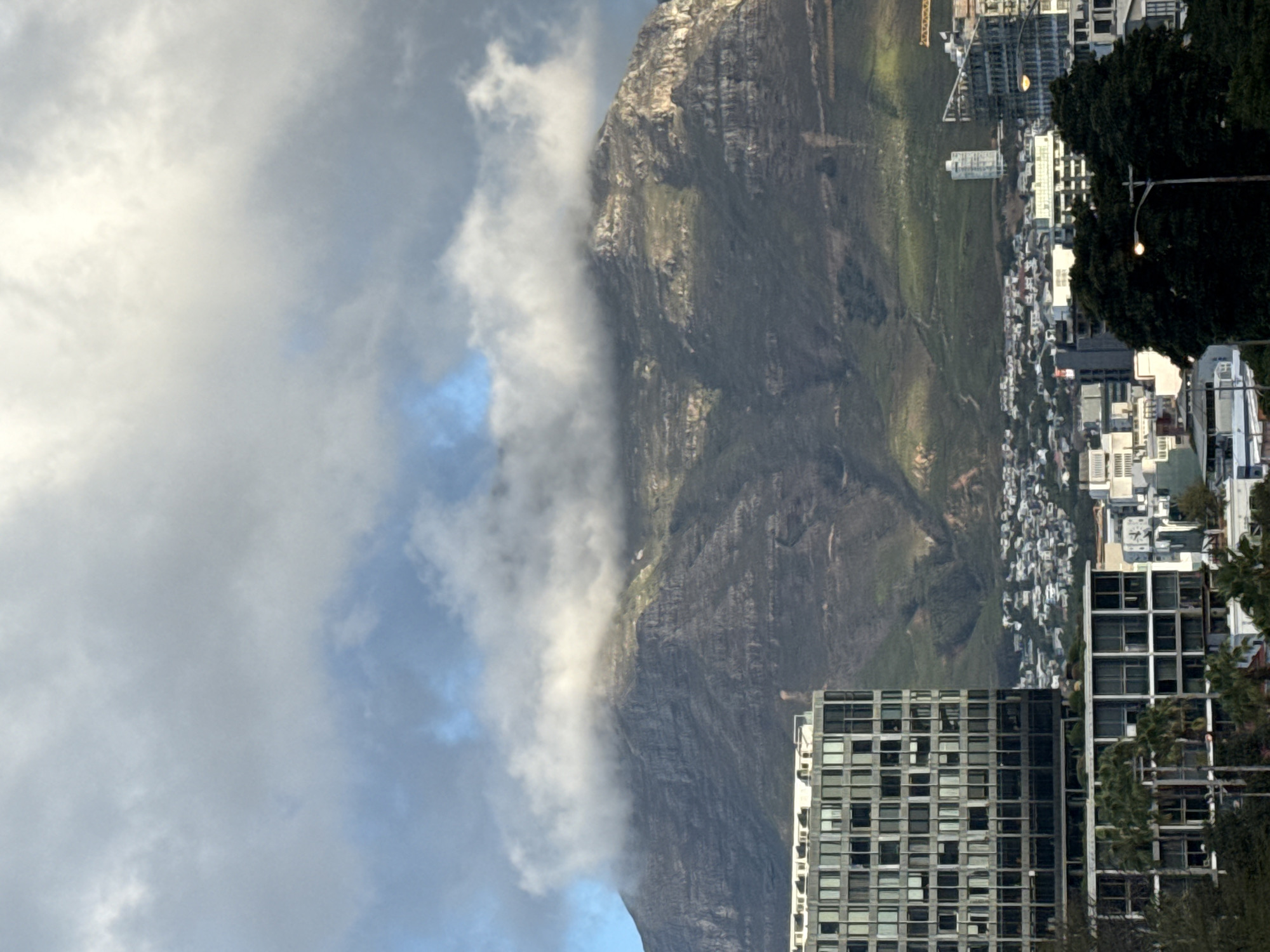 The image depicts a view in Cape Town, South Africa, featuring a mountain partially obscured by clouds on the left side. To the right of the mountain, a cityscape is visible, composed of buildings of varying heights and densities. Some high-rise buildings are prominently visible in the foreground, while residential areas can be discerned on the mountainside. Trees are present. The sky is filled with clouds. The image is rotated clockwise by approximately 90 degrees.