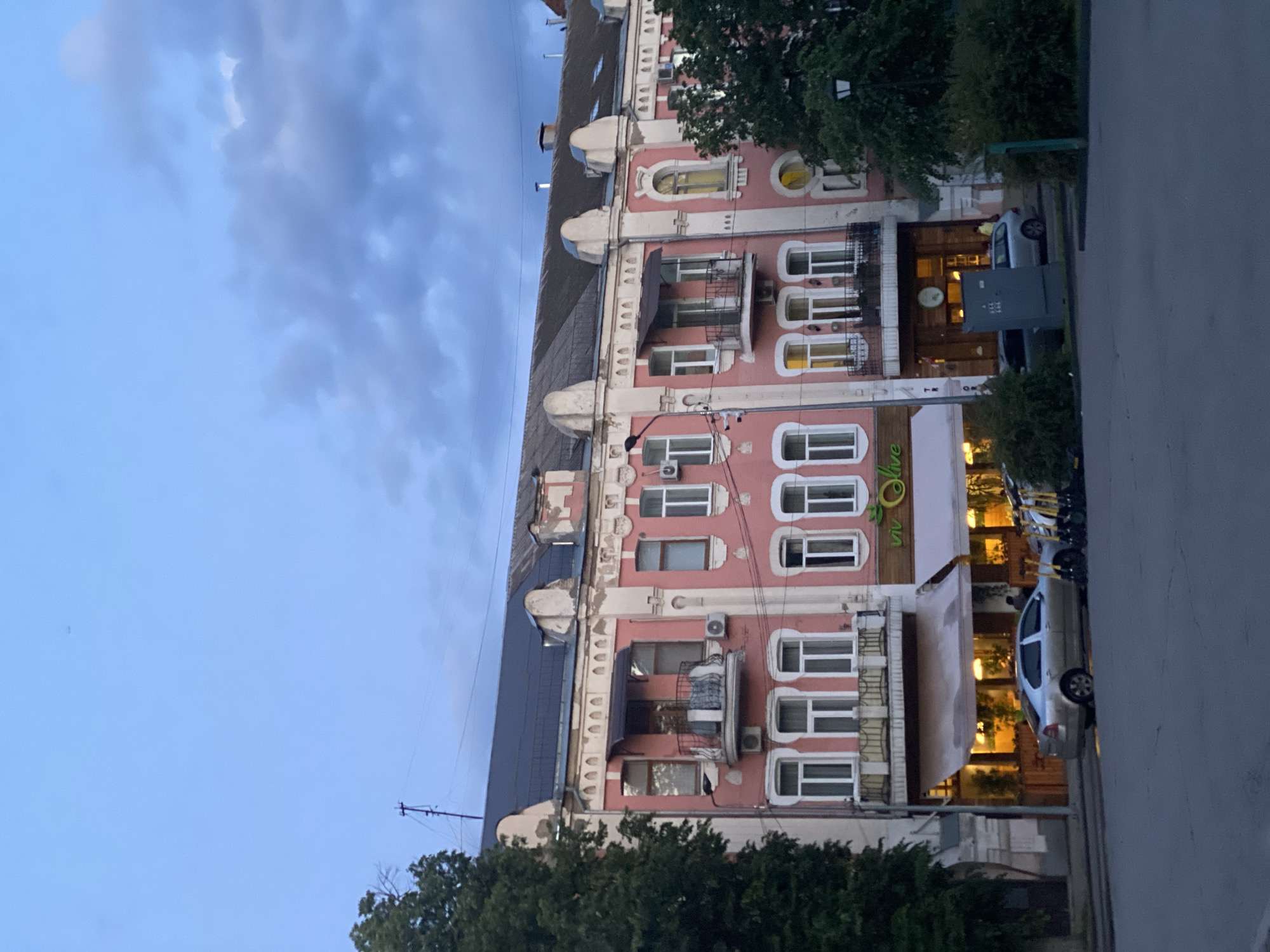 The image, originally oriented vertically, depicts a multi-story building with a distinctive pinkish-red facade and elaborate white architectural details, suggestive of an older European style. The building features a variety of window shapes, including rectangular, arched, and oval, along with several small balconies adorned with metal railings. Multiple air conditioning units are visible on the exterior walls, and the building is topped with a dark, sloped roof.
On the ground floor, a green awning clearly displays the text "Via Olive," likely indicating a restaurant or cafe. Warm, artificial lights illuminate the interior of the lower floors, suggesting it is late evening or twilight. The sky above is a soft blue with scattered, diffuse clouds.
The building fronts a paved street where several vehicles are parked, including a light-colored sedan. Green trees and bushes line the street and are scattered around the scene, partially obscuring parts of the building and a dark grey utility box on the sidewalk. A streetlight pole is also visible. The overall scene is a calm street view in Poltava, Ukraine, during the evening. No specific human activity is prominent.