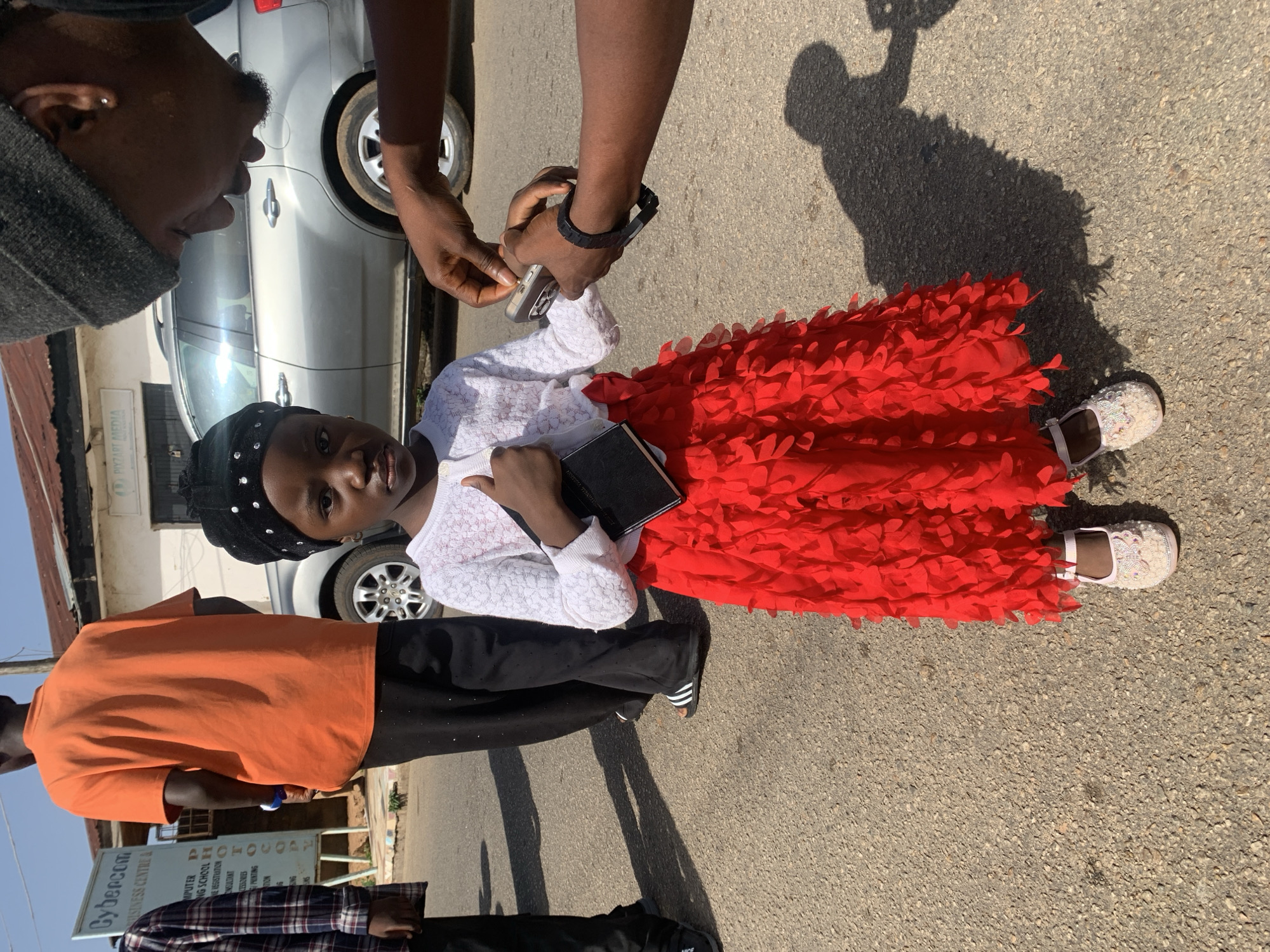 A young girl in a red frilled skirt and white top stands on a paved surface. She wears black leggings or pants and white shoes. She also wears a black head covering with white adornments. The girl is holding a black book. An individual is holding a phone in their hand, possibly taking a photo.
Behind the girl, there is a partially visible silver vehicle. Other people are visible, one wearing an orange shirt and another a plaid shirt. The background includes a building and a sign that reads, in part, "Cybercom BUSINESS CENTRE & COMPUTER NING SCHOOL". The sign indicates “PIYZART MEDIA” and likely mentions “Jos Nigeria”. The presence of Cybercom BUSINESS CENTRE & COMPUTER likely places this photo in Jos, Nigeria.