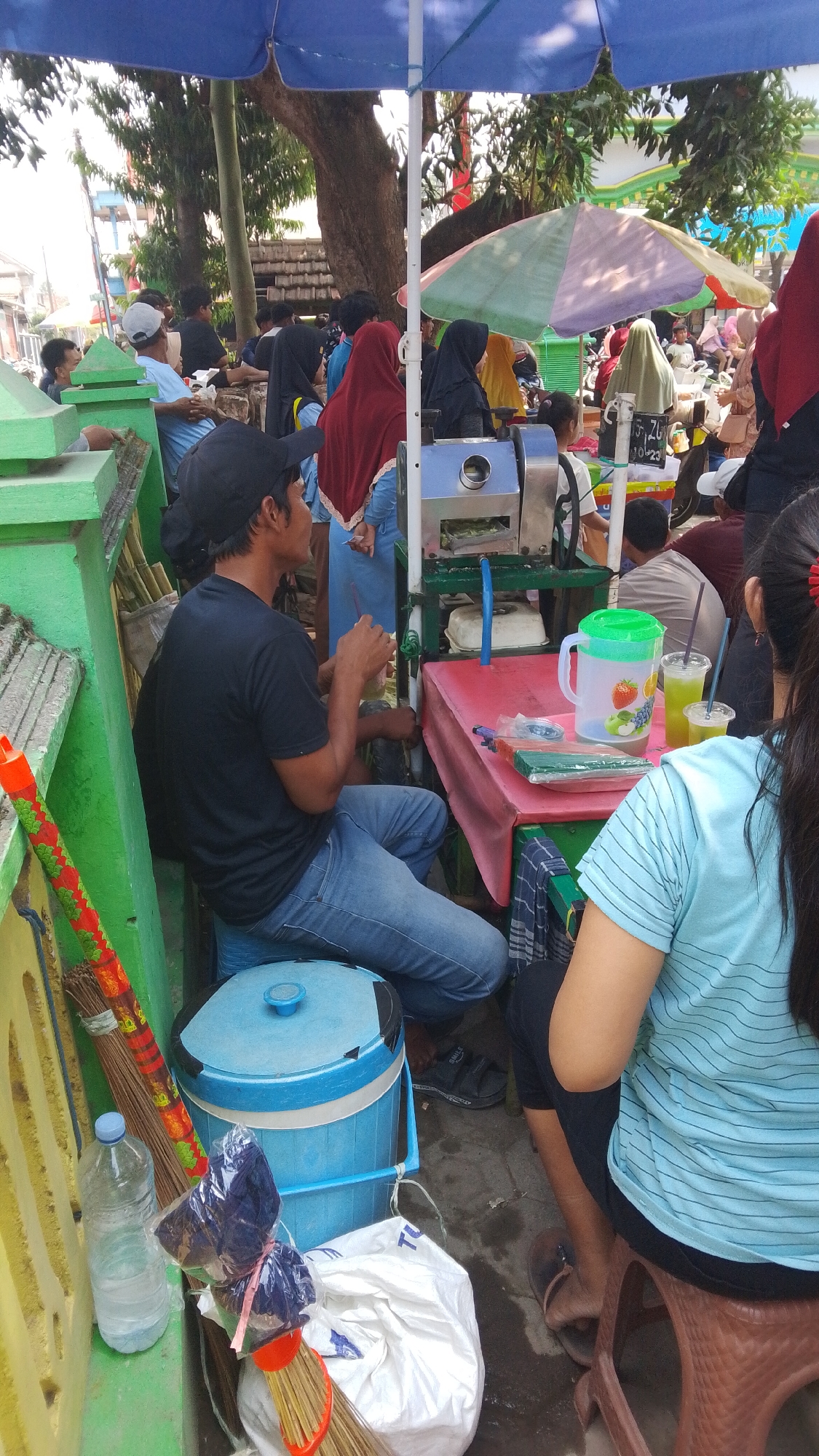 In Mancar, Indonesia, a street vendor operates a sugar cane juice stand under a blue tarp. A man in a black t-shirt and blue jeans sits on a blue cooler, holding a straw in a glass of juice. In front of him, a red table displays a sugarcane juicing machine, a pitcher with fruit illustrations, and two plastic cups of juice. To the left, a broom leans against a green fence. Several customers are visible in the background, some wearing hijabs. The scene suggests a typical outdoor market or roadside gathering.