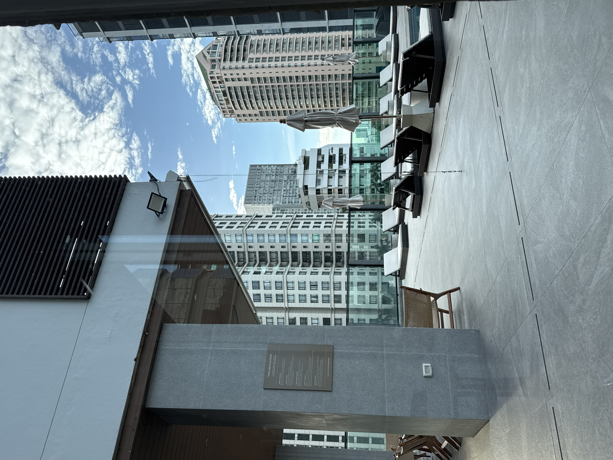 This is a photograph of a high-rise building's rooftop pool area in Bangkok, Thailand. The foreground shows a tiled patio with several lounge chairs and umbrellas. A large grey pillar with a sign displaying "POOL REGULATIONS" stands to the left. In the background, modern skyscrapers dominate the skyline under a partly cloudy blue sky. The architecture suggests a contemporary urban setting. There are no people visible in the image.