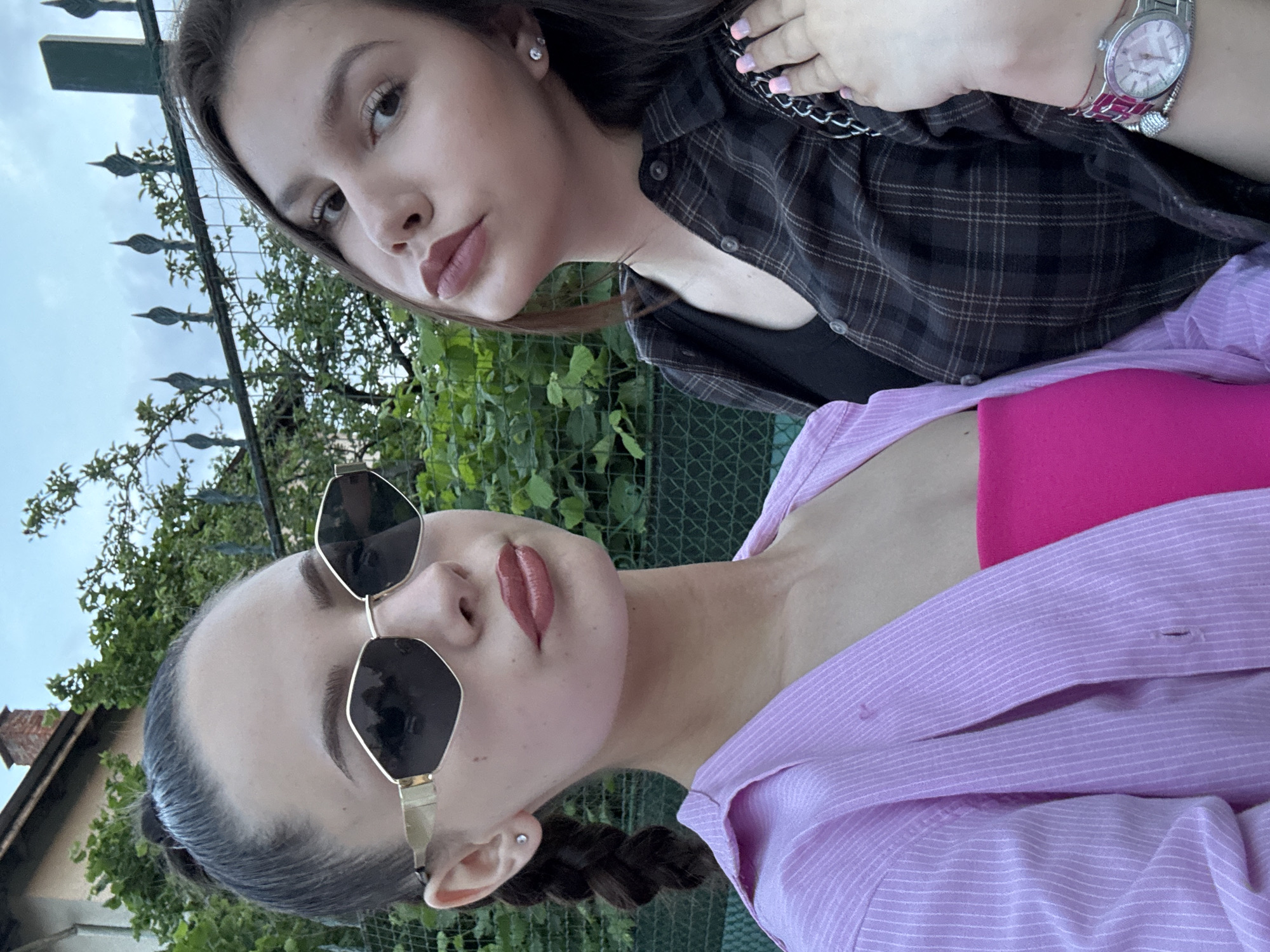 Two young women are captured in a close-up, eye-level outdoor image. The woman on the left, with dark hair styled in a high bun and a visible braid on her left side, wears large hexagonal gold-rimmed sunglasses with dark lenses. Her lips are coated with pink-mauve lipstick, and a silver stud earring is visible in her left earlobe. She is dressed in a light pink, vertically striped, collared shirt worn open over a bright fuchsia strapless top. The woman on the right, positioned slightly behind and to the right, has long dark hair parted on the side. She wears a dark brown plaid button-up shirt over a black top and also has pink-mauve lipstick; a silver stud earring is visible in her right ear. Her left hand, with light pink nail polish, rests near her chest, displaying a silver chain strap, possibly from a bag. A silver watch with a pink strap and a beaded bracelet are visible on her right wrist. The background features dense green foliage, a dark green metal fence with decorative spires, and a partial view of a light-colored building with a brick chimney against an overcast sky. A green mesh fence is visible in the lower background.